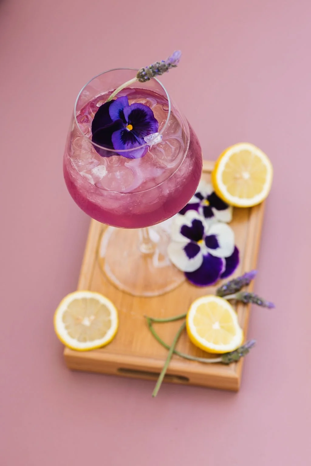 A pink cocktail garnished with a purple edible flower and lavender, served on a wooden tray along with lemon slices and purple and white flowers, on a pink background.