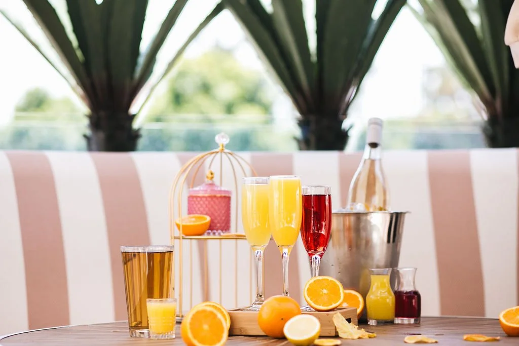 Assorted beverages including orange juice, champagne, and wine on a table with oranges and lemon slices, with a bottle of wine in an ice bucket in the background.