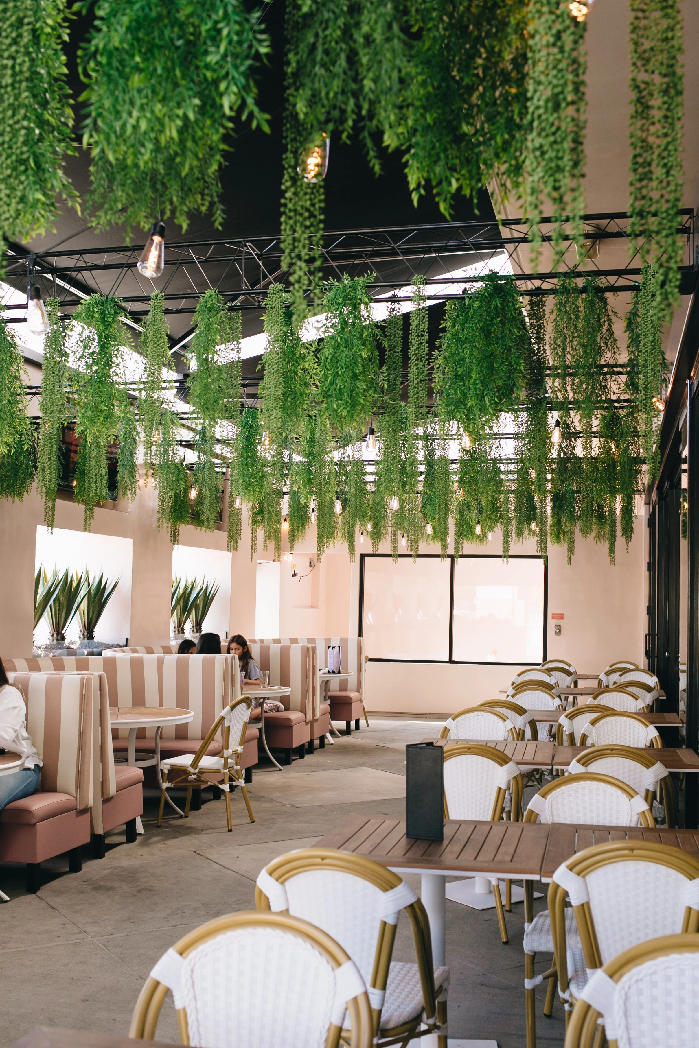 Interior of a cafe with hanging greenery and string lights on the ceiling, beige upholstered booths along the wall, and white chairs with gold accents around wooden tables.
