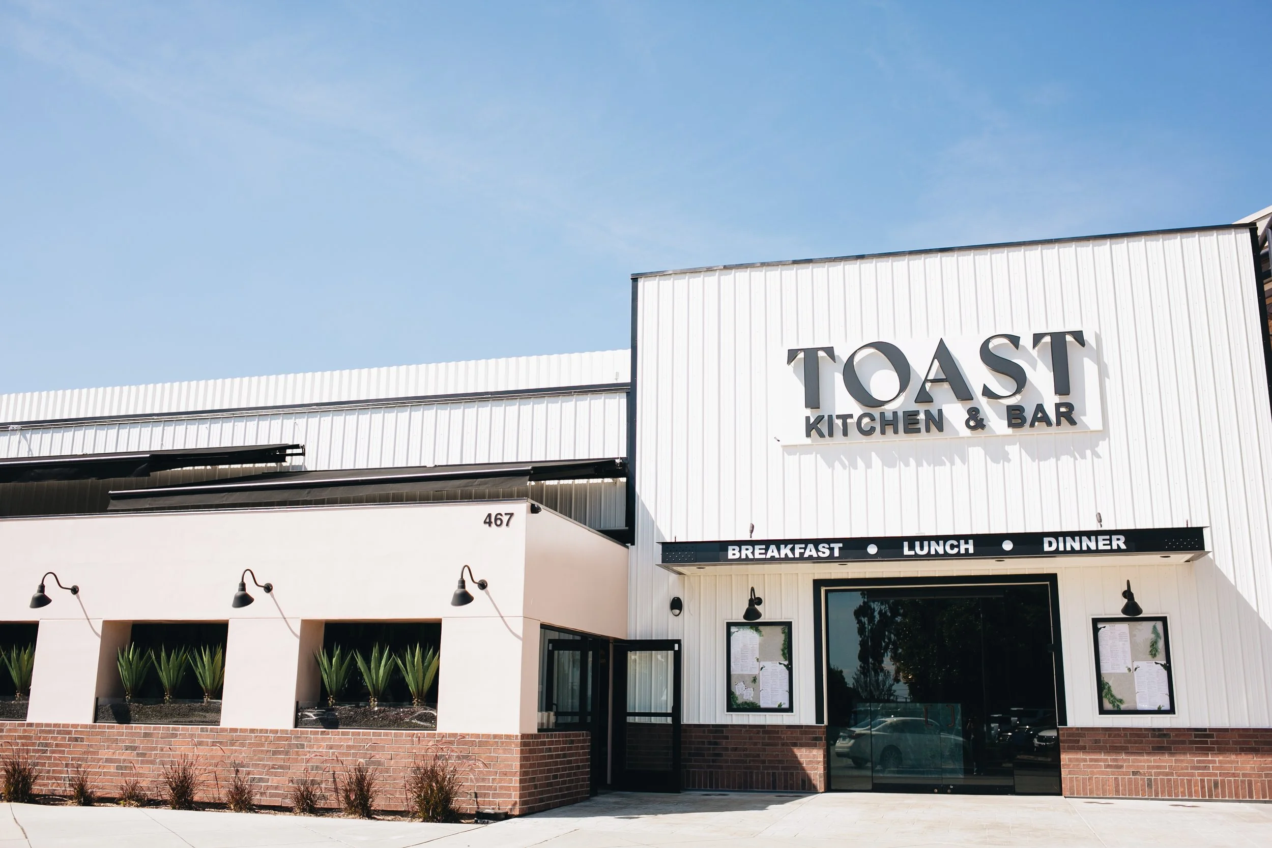 Exterior view of Toast Kitchen & Bar restaurant with white building, black signage, and glass entrance, under a clear blue sky.