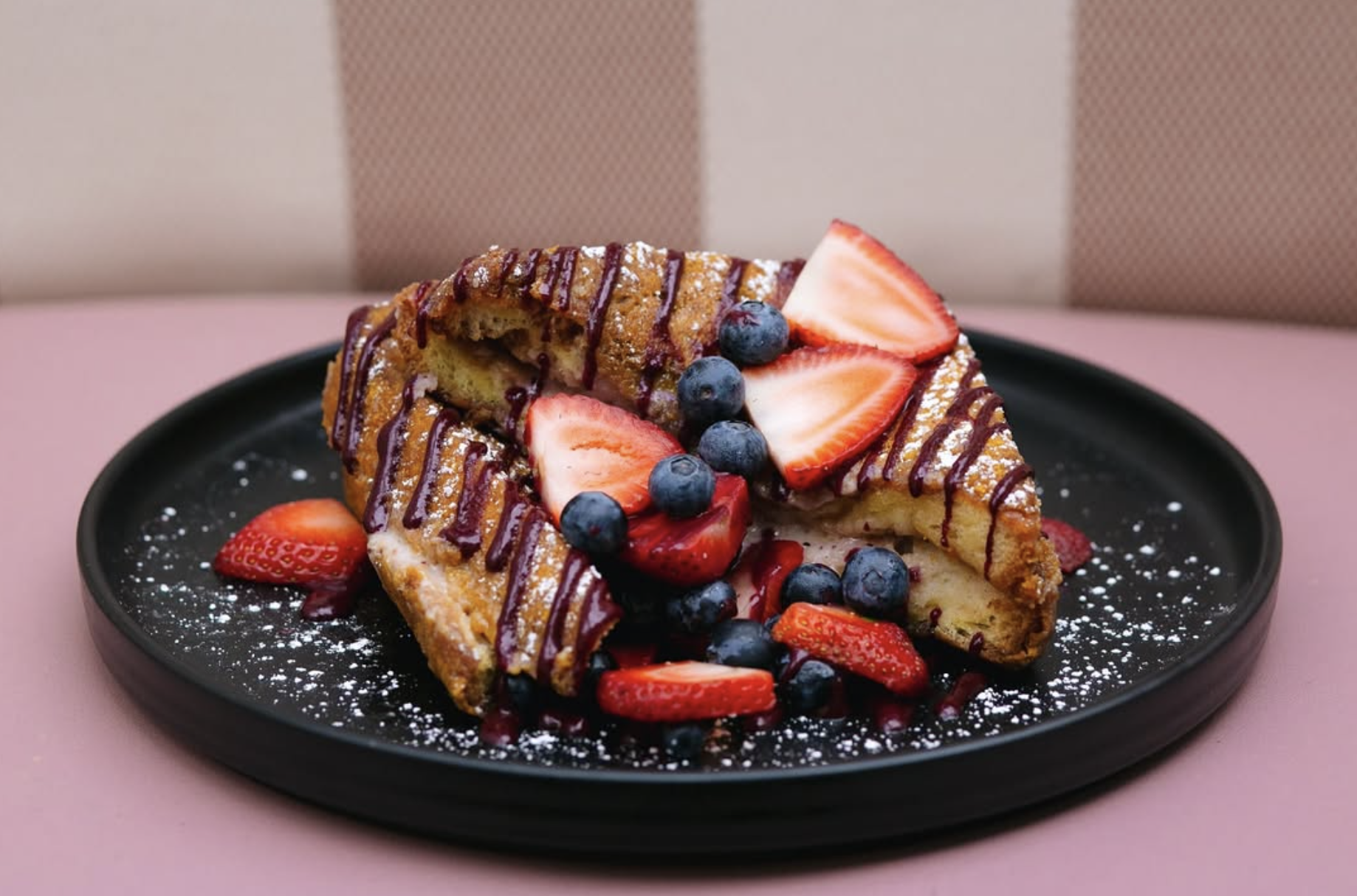 A serving of French toast topped with fresh strawberries, blueberries, and drizzled chocolate sauce on a black plate with powdered sugar, placed on a pink table.