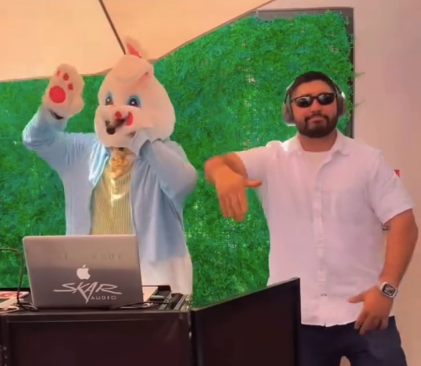 Person wearing a rabbit costume with a DJ booth next to a man in a white shirt and sunglasses, both striking a pose in front of a green background.