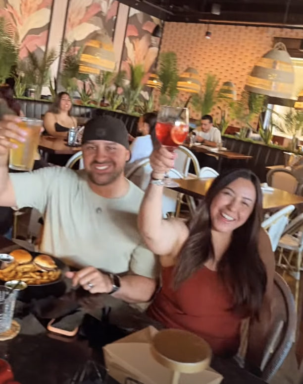 Two people celebrating at a restaurant, holding drinks and smiling. The man wears a black bandana and a light-colored shirt, while the woman has long dark hair and a reddish-brown top. There are glasses of drinks, a plate of burgers and fries on the table, and other diners in the background. The restaurant has tropical-themed decor with plants, colorful mural, and hanging lights.