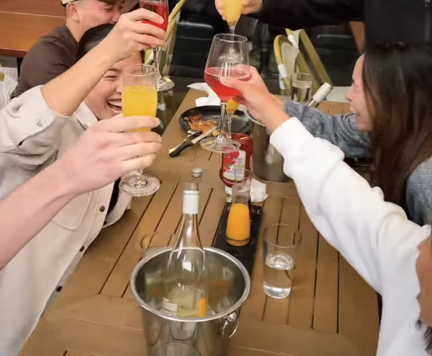 Group of friends at a dinner table raising glasses for a toast, with drinks and condiments present.