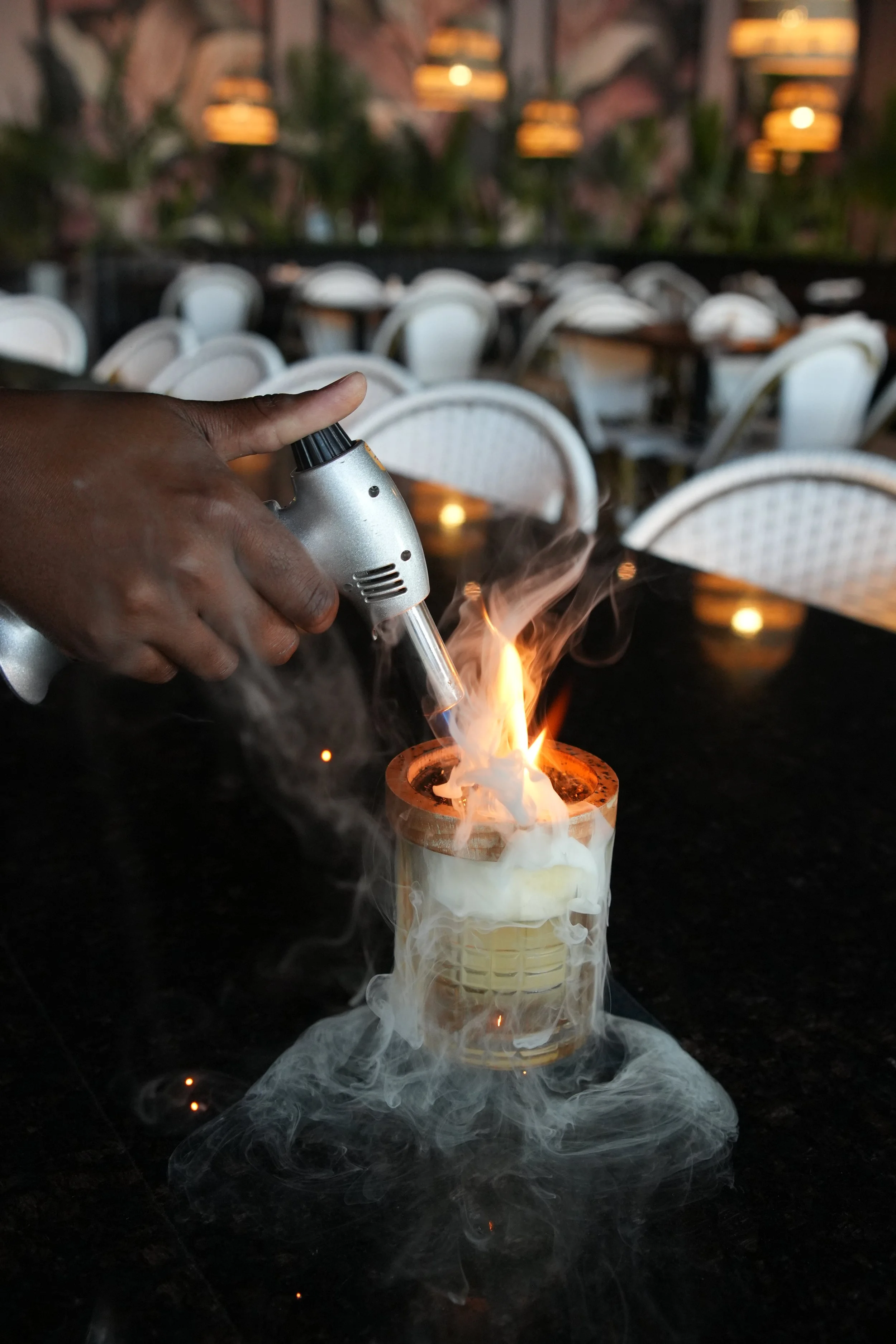 A person using a small blowtorch to ignite a substance in a glass container on a table in a restaurant or cafe with empty chairs and tables, and warm hanging lights in the background.