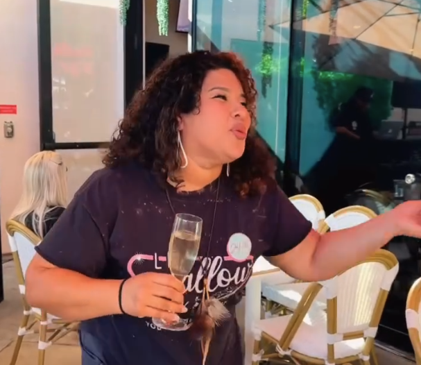 A woman with curly hair holding a glass of champagne at a celebration or party, smiling and gesturing with her hand.