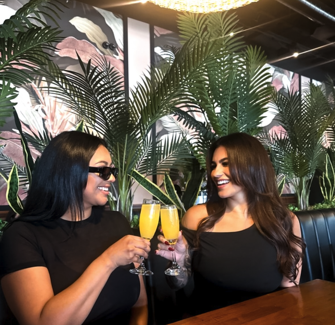 Two women celebrating with glasses of yellow drinks at a restaurant, surrounded by lush green plants and tropical-themed artwork on the wall.