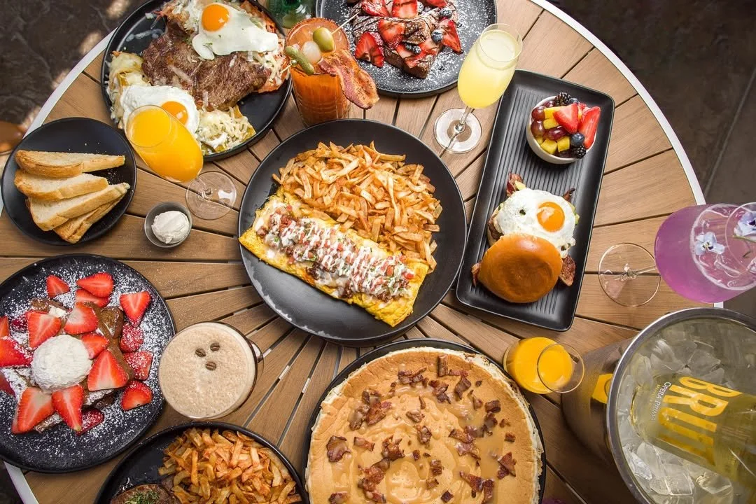 A variety of breakfast foods on a round wooden table, including eggs, bacon, toast, fries, pancakes with strawberries and powdered sugar, fruit bowl, and drinks like orange juice and mimosas.
