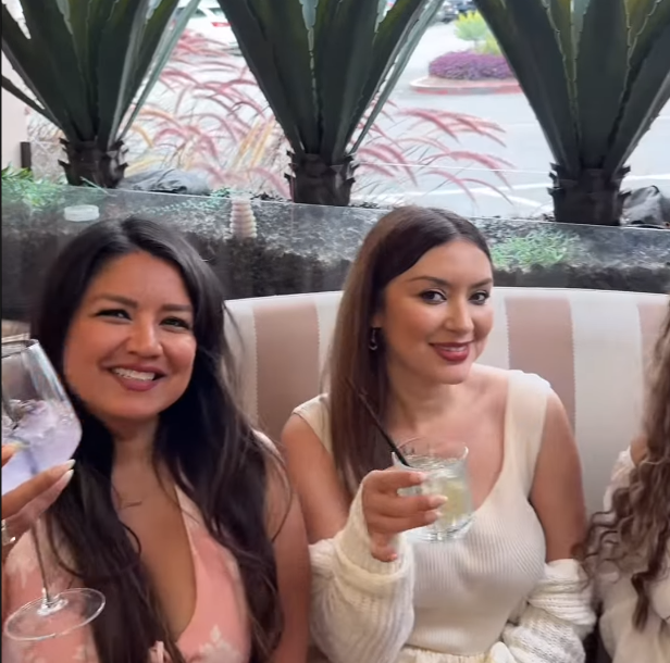 Two women sitting at a restaurant table, smiling and holding drinks, with a background of large potted plants.