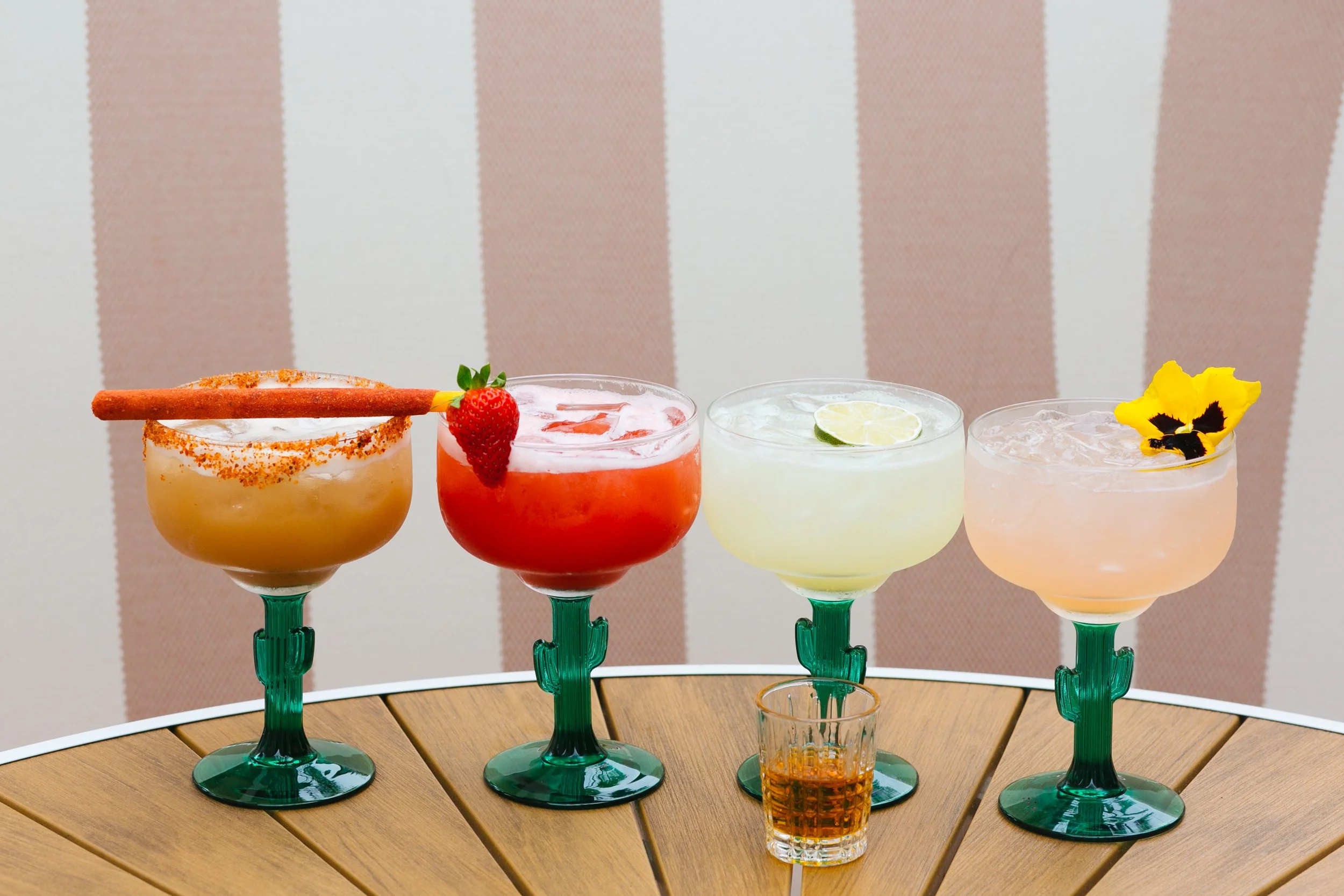 Four colorful cocktails with garnishes and a small shot glass on a wooden table.