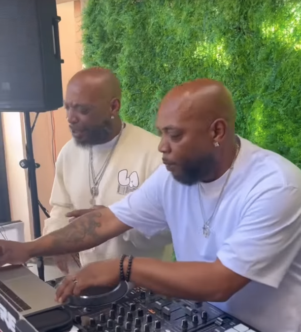 Two men with shaved heads and beards, one wearing a white t-shirt and the other a cream-colored shirt, are at a DJ mixing console with a laptop, in front of a green leafy wall.