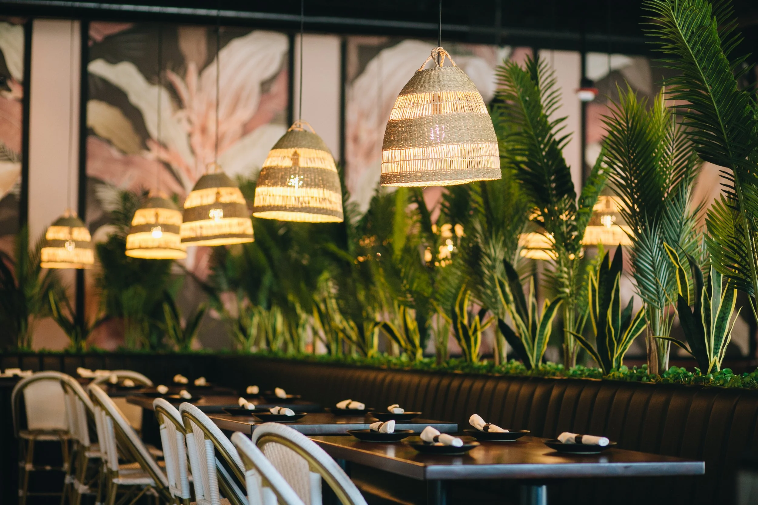 Restaurant interior with hanging wicker pendant lights, tropical plants, and a long dining table set with black plates and white napkins.