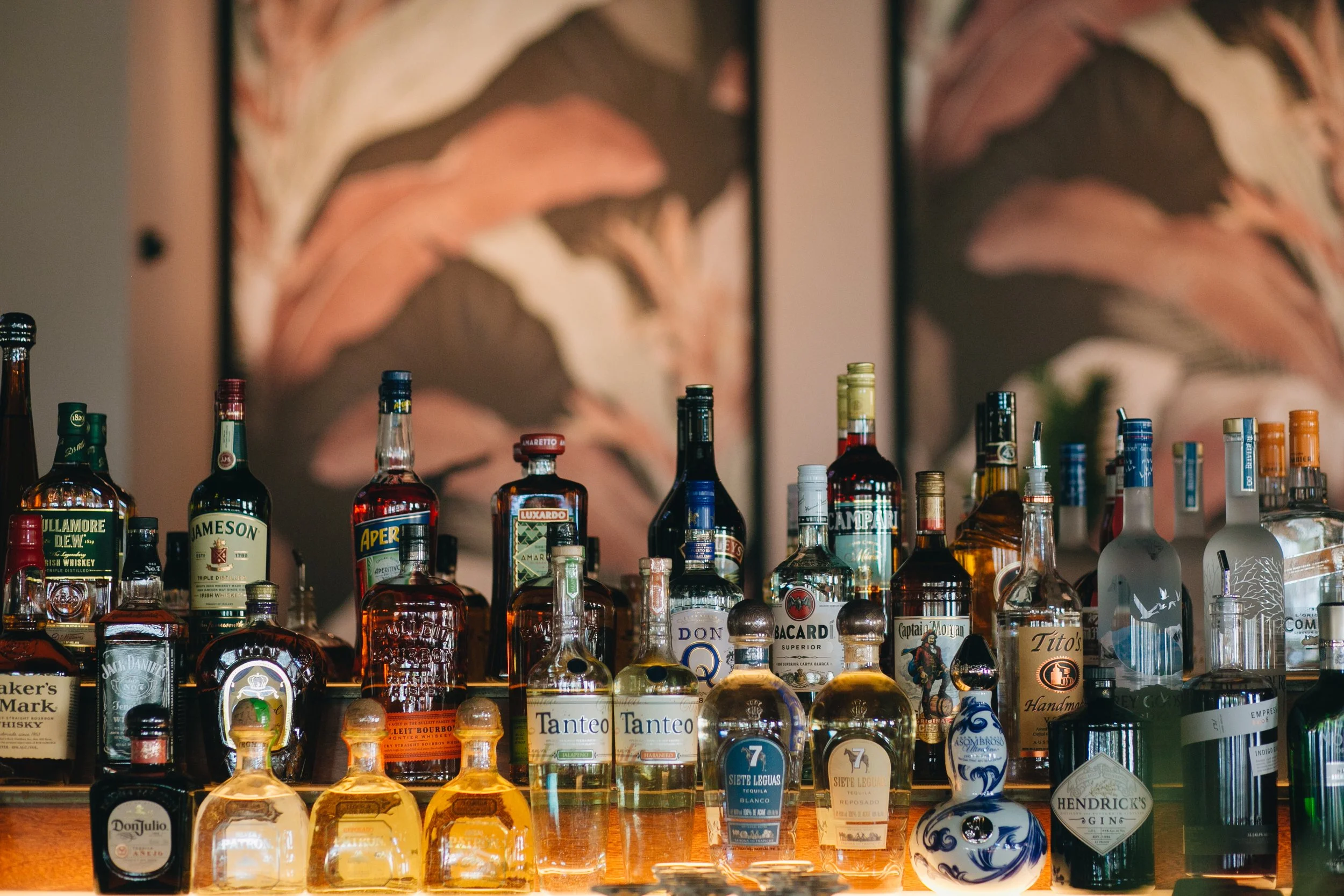 Assorted liquor bottles on a bar shelf with colorful abstract artwork in the background.