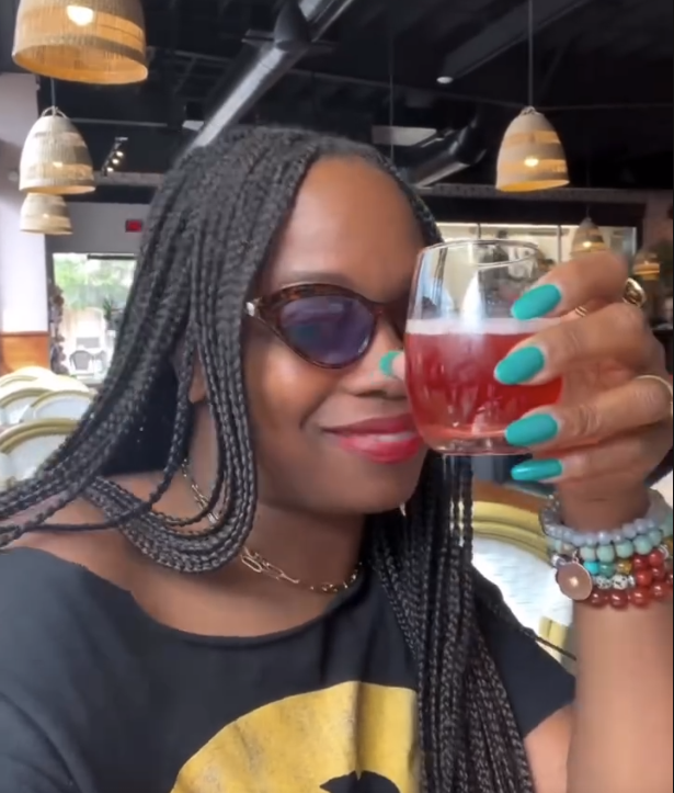 A woman with braids wearing sunglasses, holding a glass of red-colored drink, smiling inside a well-lit restaurant.