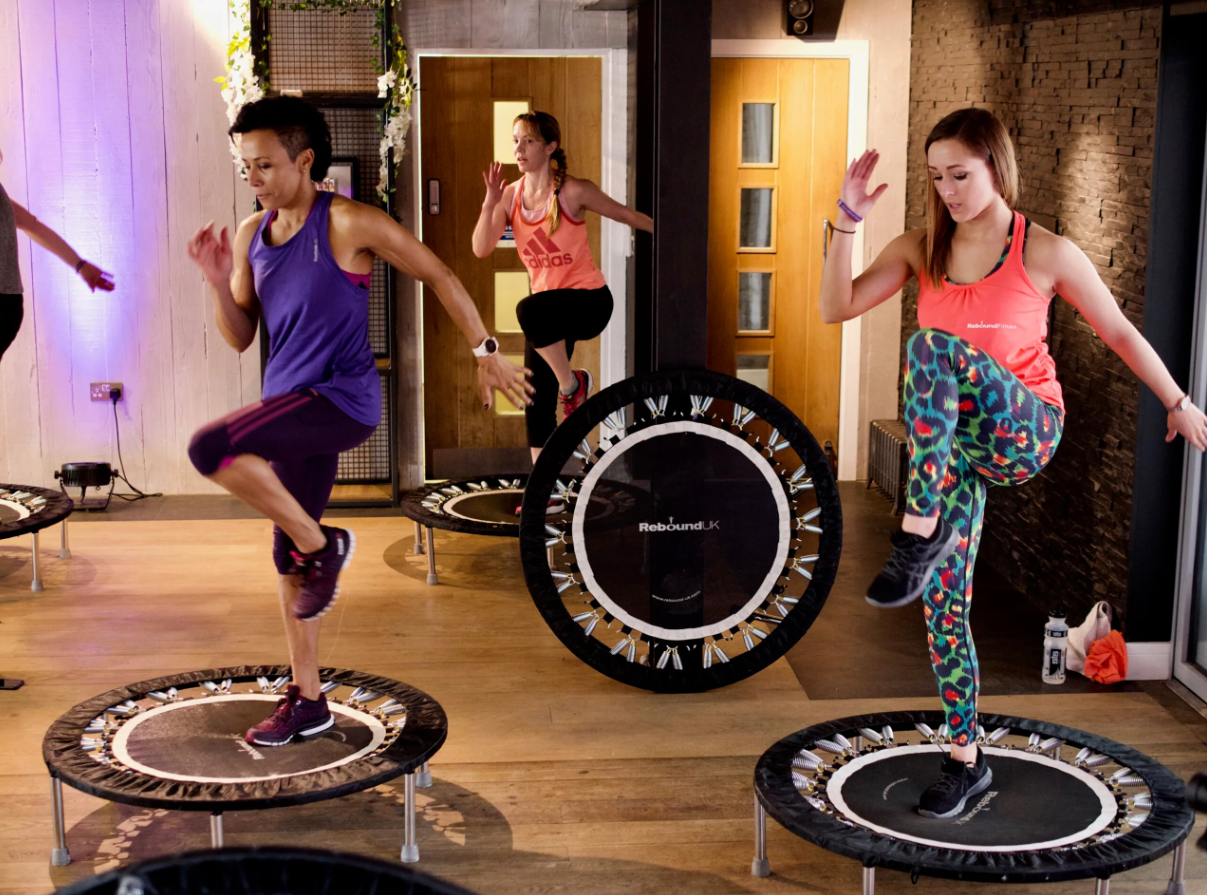 Three women participating in a trampoline fitness class indoors, jumping on mini trampolines.