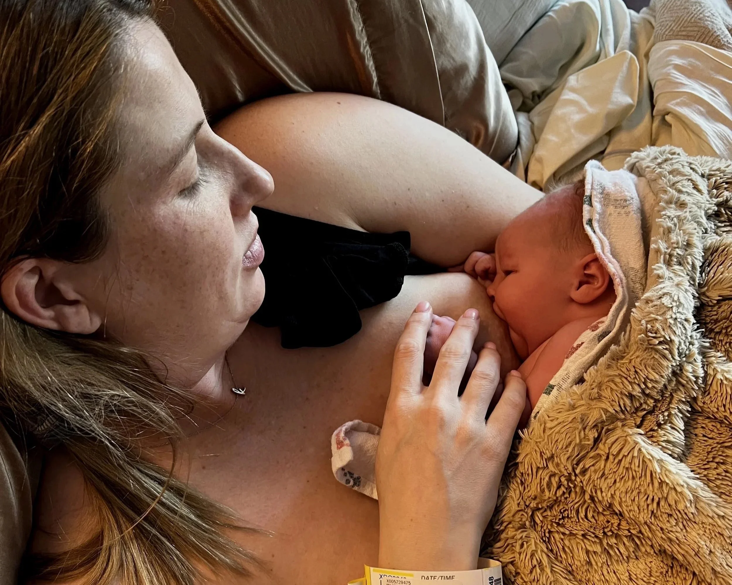 A woman with red hair is lying down on a bed, breastfeeding a newborn baby who is wrapped in a blanket. The woman has her eyes closed and is touching the baby gently.