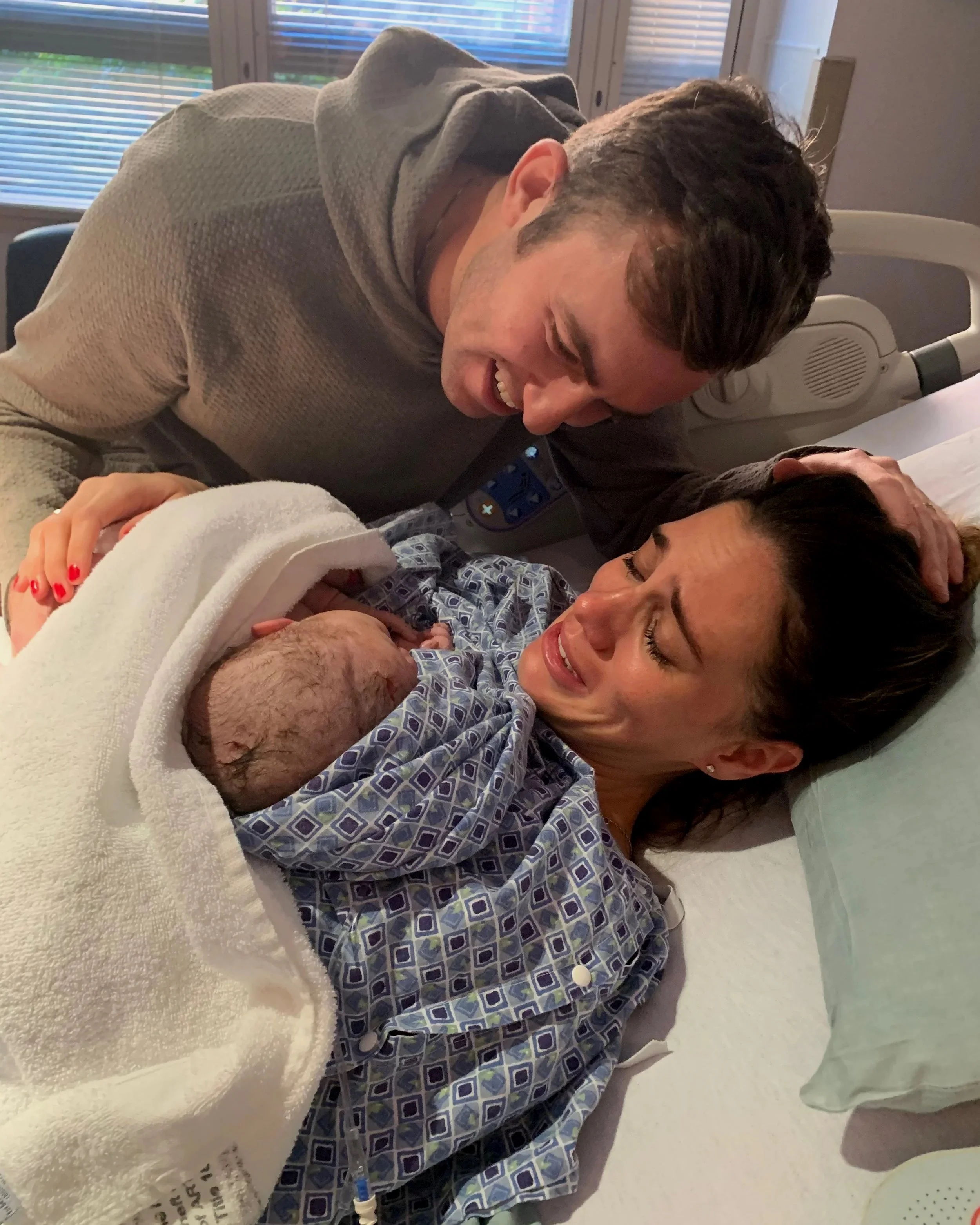 A woman lying in a hospital bed holding a newborn baby, with a man leaning over her smiling and touching her head.
