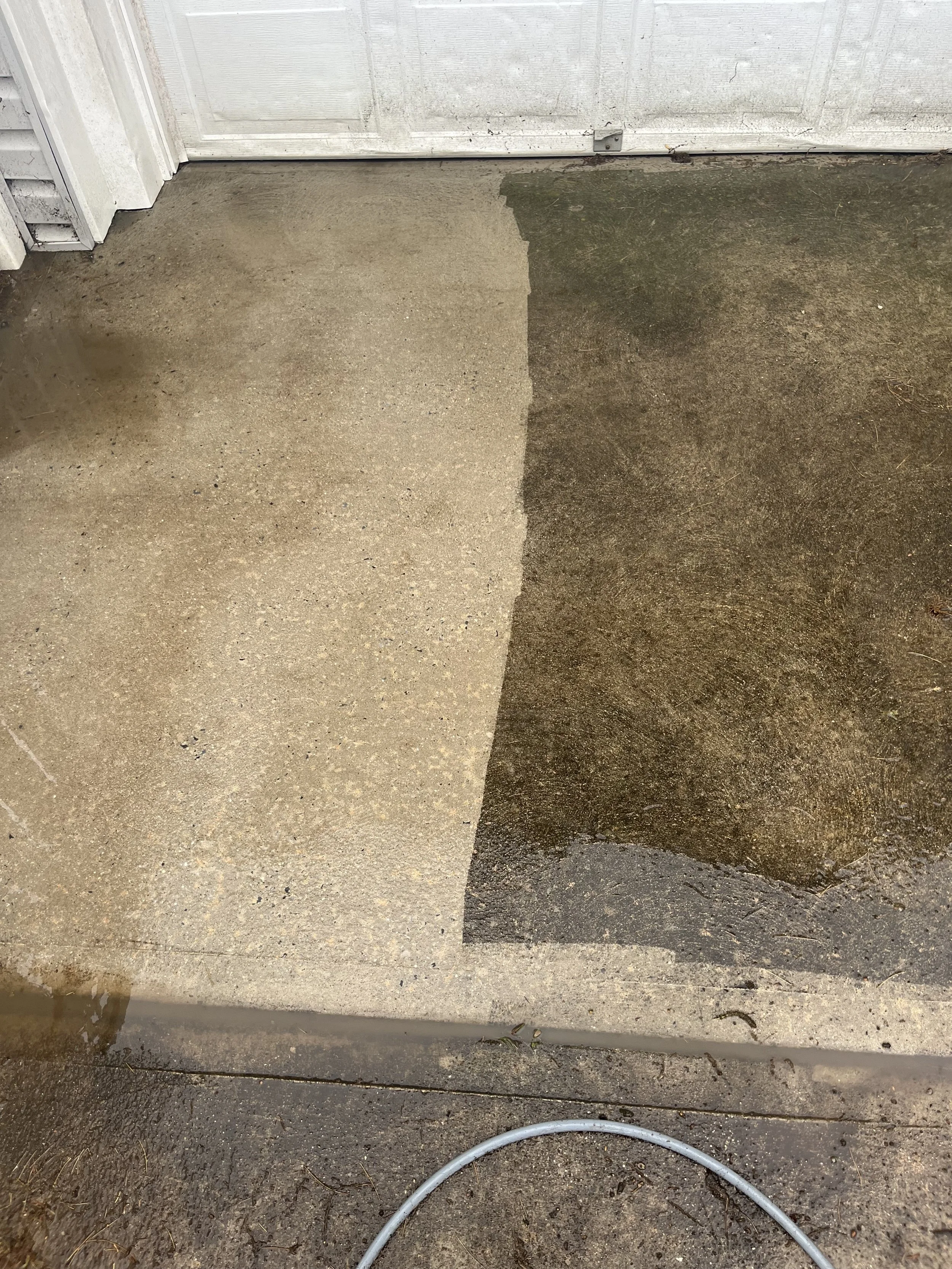 Sidewalk and driveway area with contrasting concrete sections, wet and dry areas, and a garden hose on the ground near a garage door.