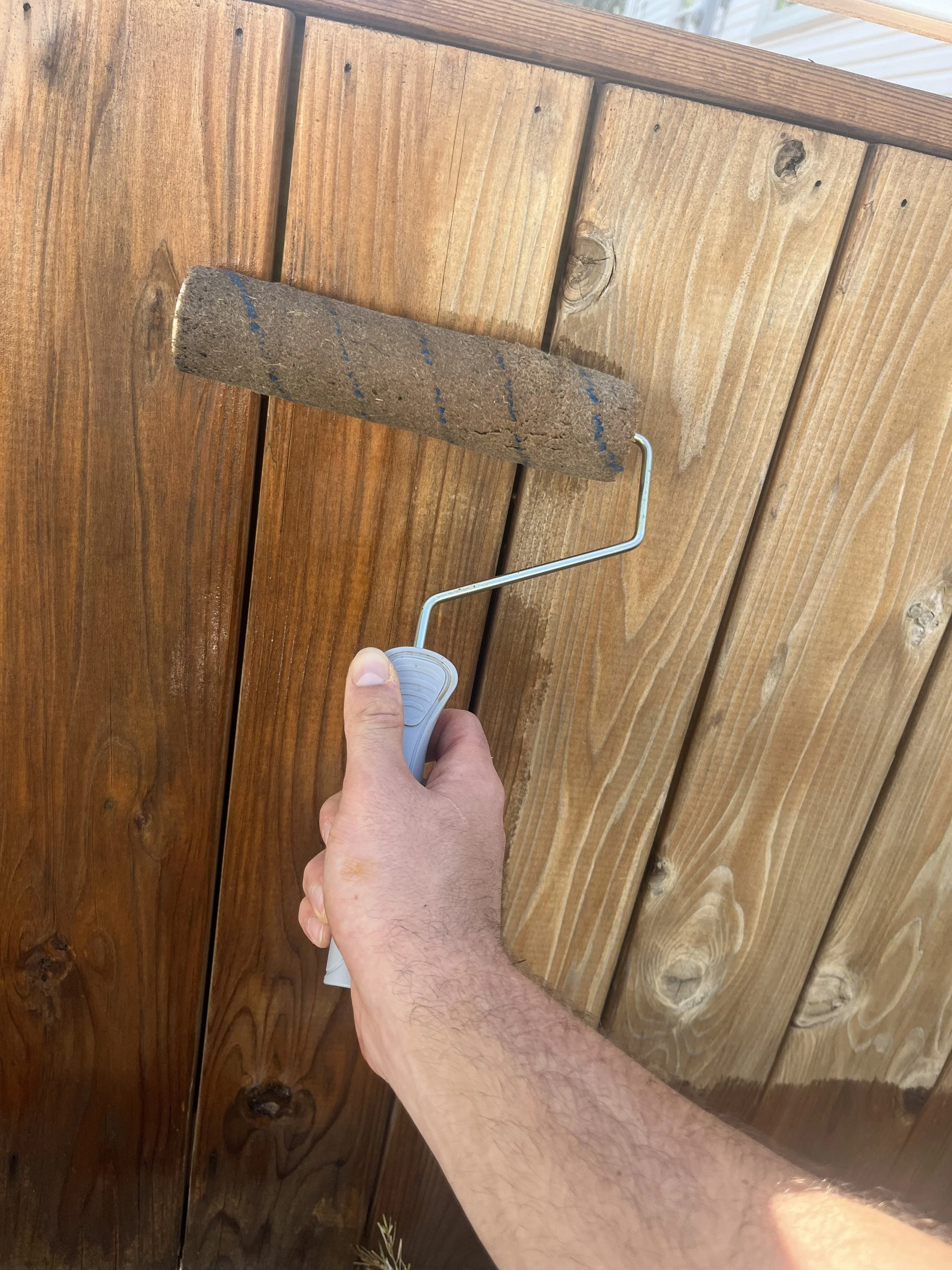 A person holding a paint roller with a brown, textured roller cover and a metal handle, painting a wooden fence with vertical planks.