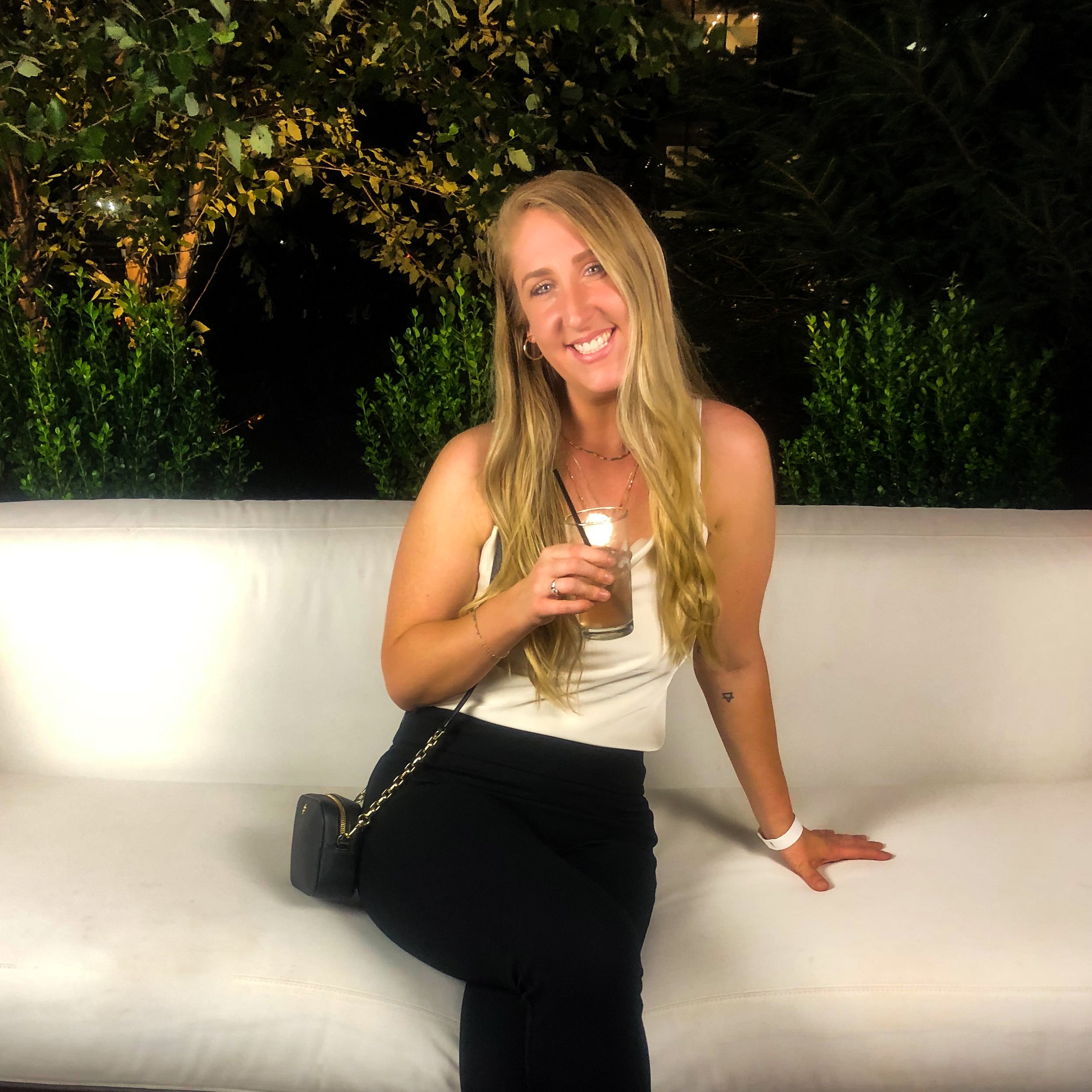 A woman with long blonde hair sitting on a white outdoor couch, holding a drink, smiling at the camera, with greenery in the background at night.