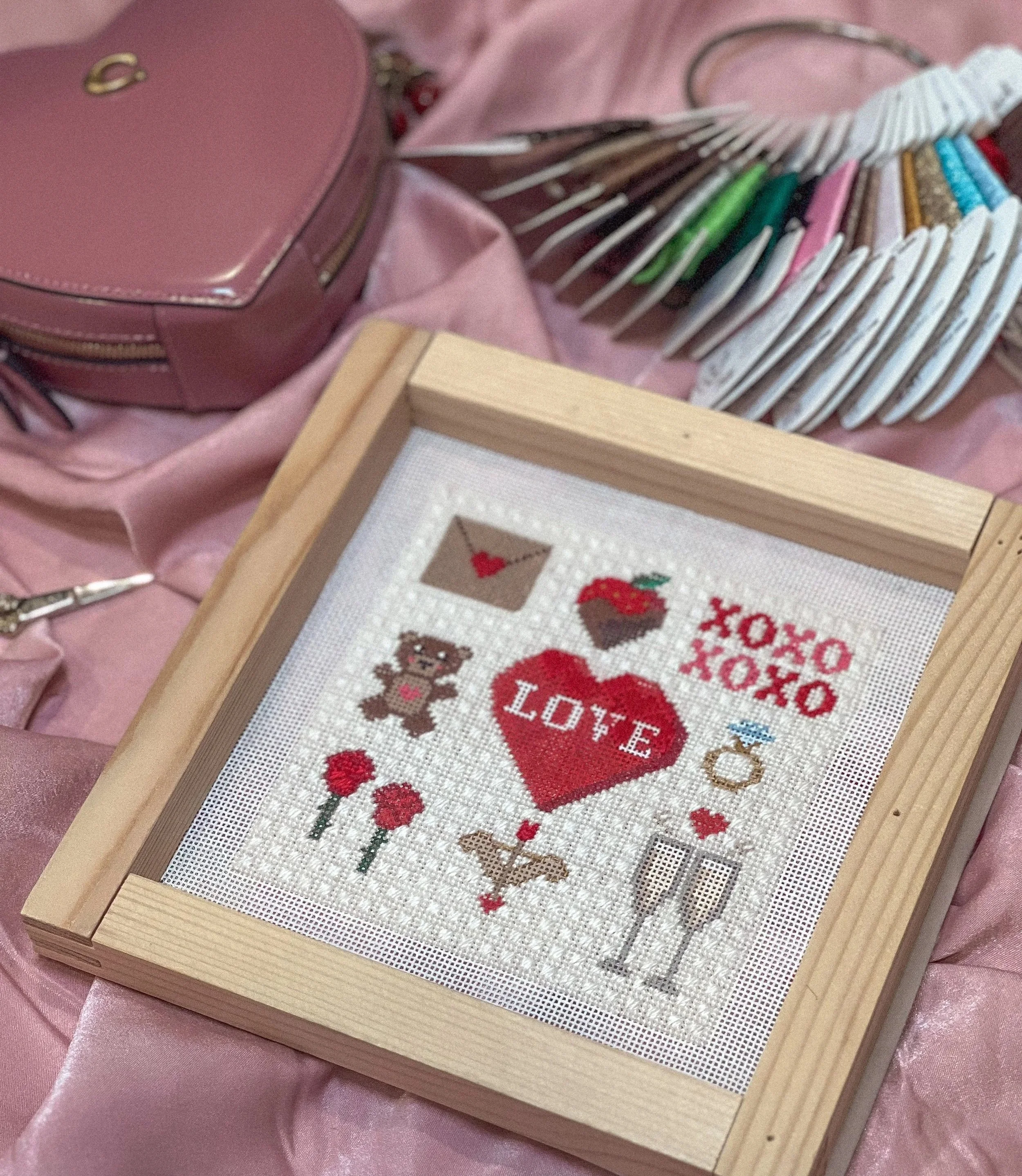 Needlepoint canvas with love-themed designs including a heart with 'LOVE' inside, a teddy bear, flowers, champagne glasses, a ring, and the words 'XOXO' and 'XO' on a white fabric background, framed in a light wood frame.
