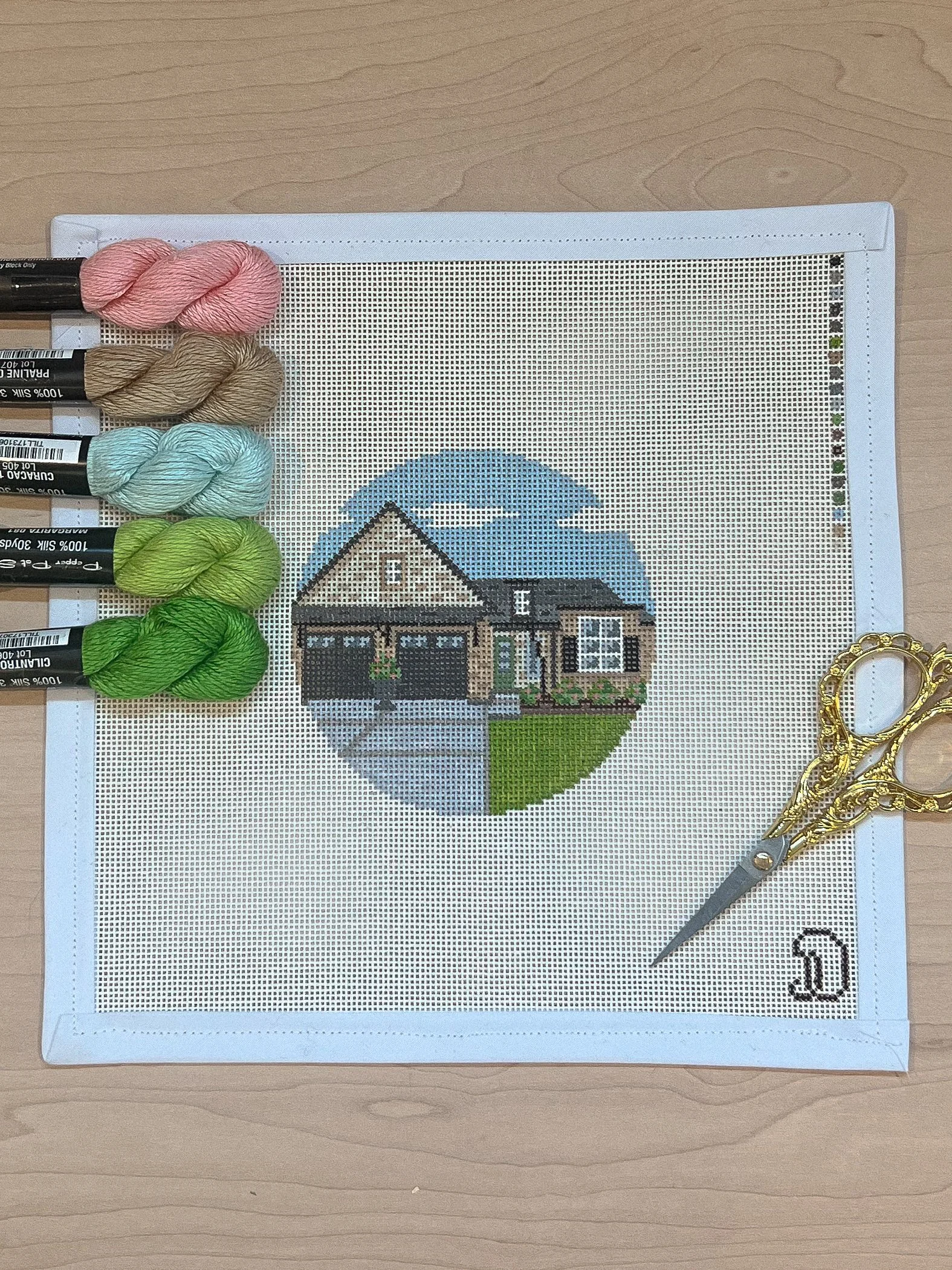 Needlepoint canvas with a house design, threads, and scissors on a wooden surface.