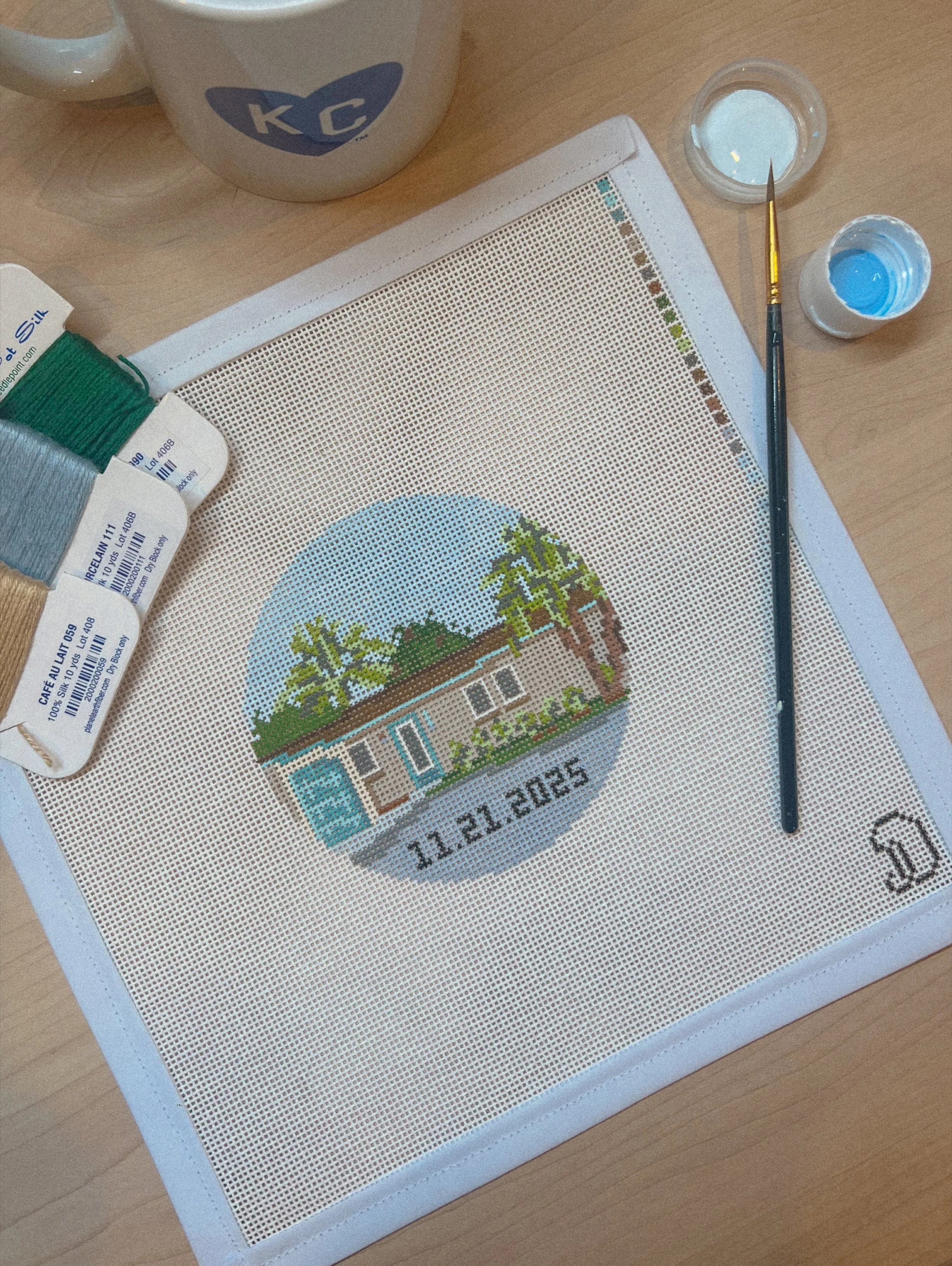 Needlepoint canvas with a house surrounded by trees, the date 11.21.2025 stitched below, and a small horse outline in the corner, on a wooden table with paint, brushes, and thread.