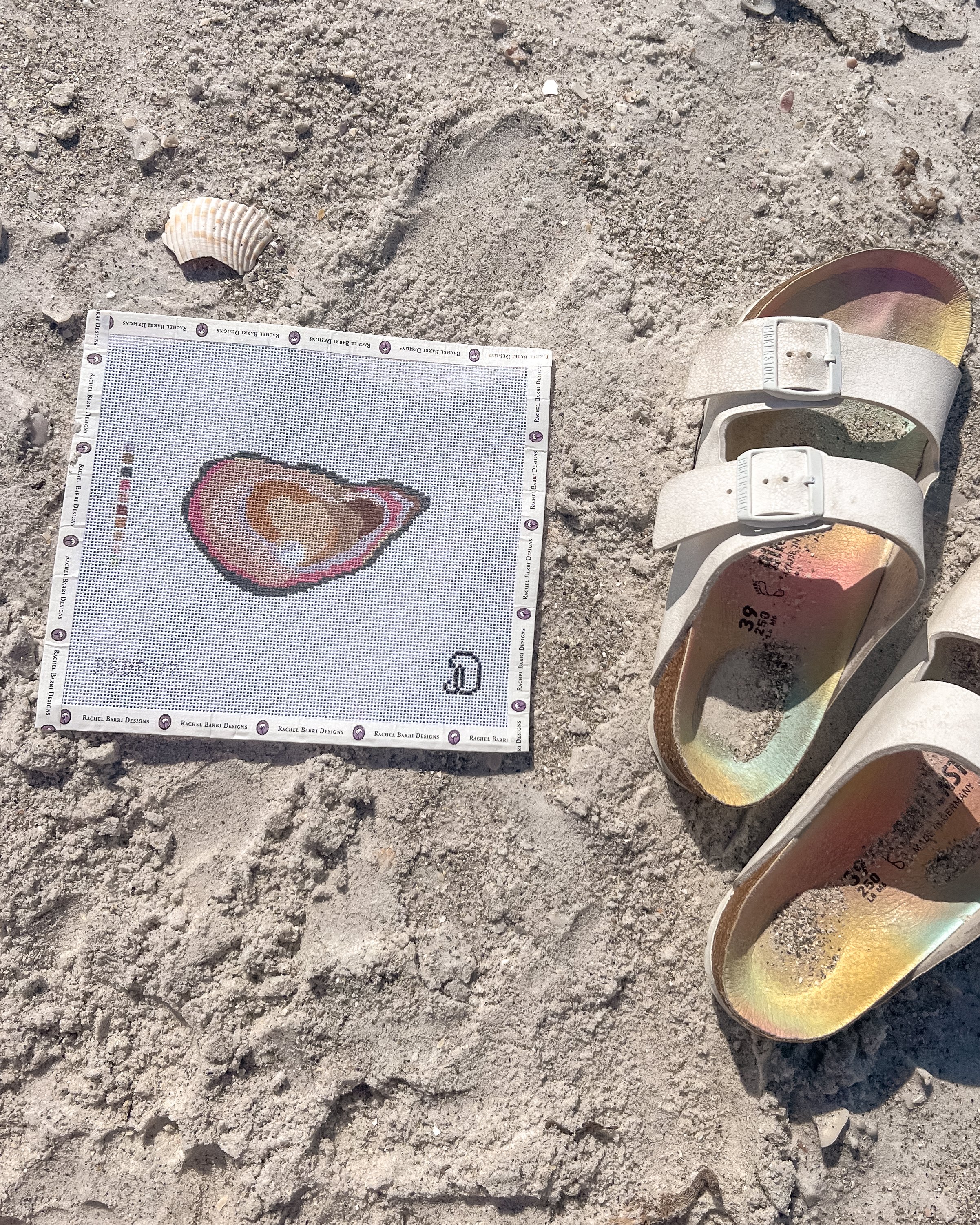 Needlepoint design of a pink oyster shell, placed on sandy beach ground next to a seashell and white sandals with sand inside.