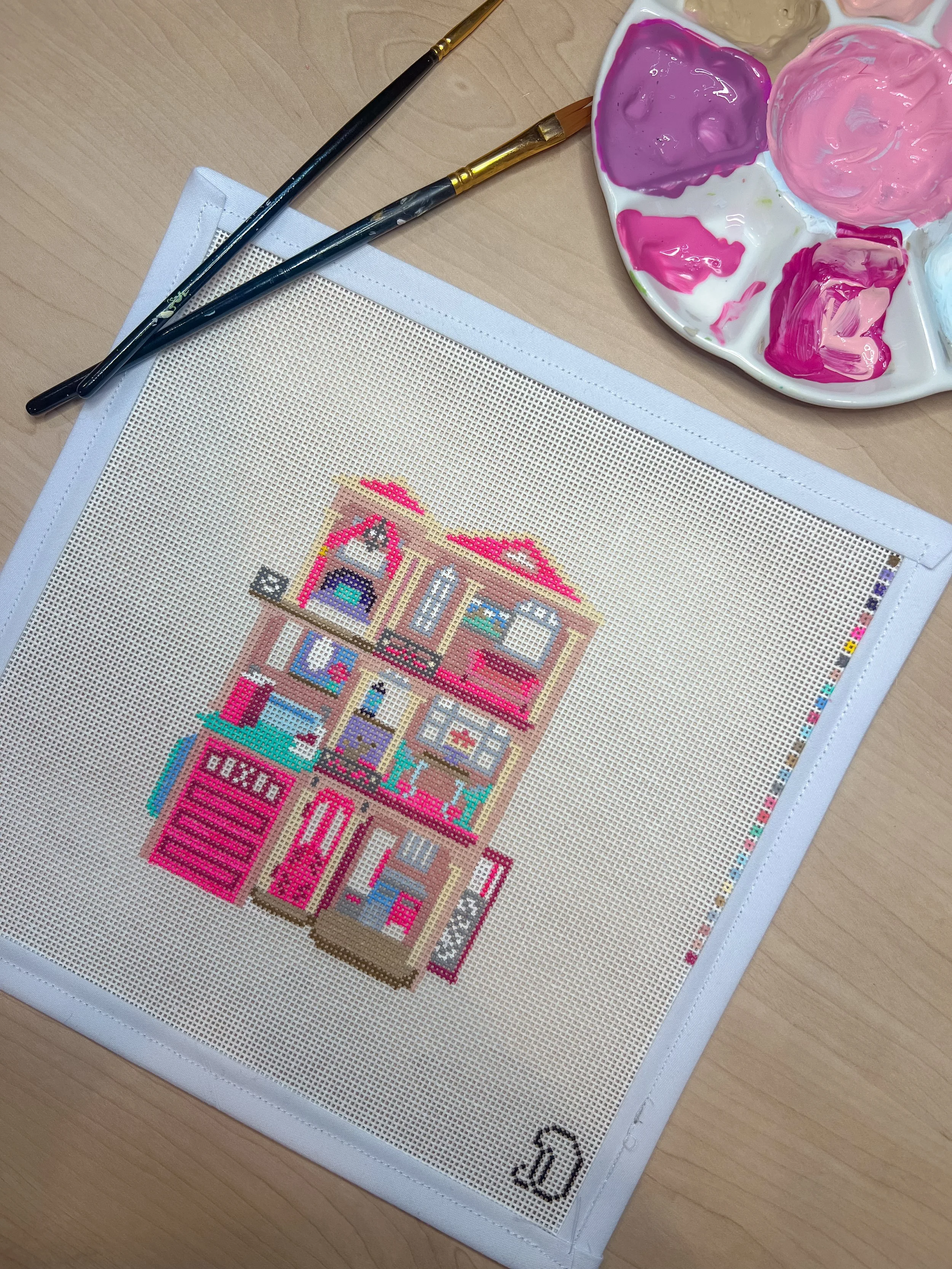 Needlepoint canvas painted with colorful toy dollhouse Barbie dream house on a white mesh canvas, on a wooden table with a paint palette and two paintbrushes nearby.
