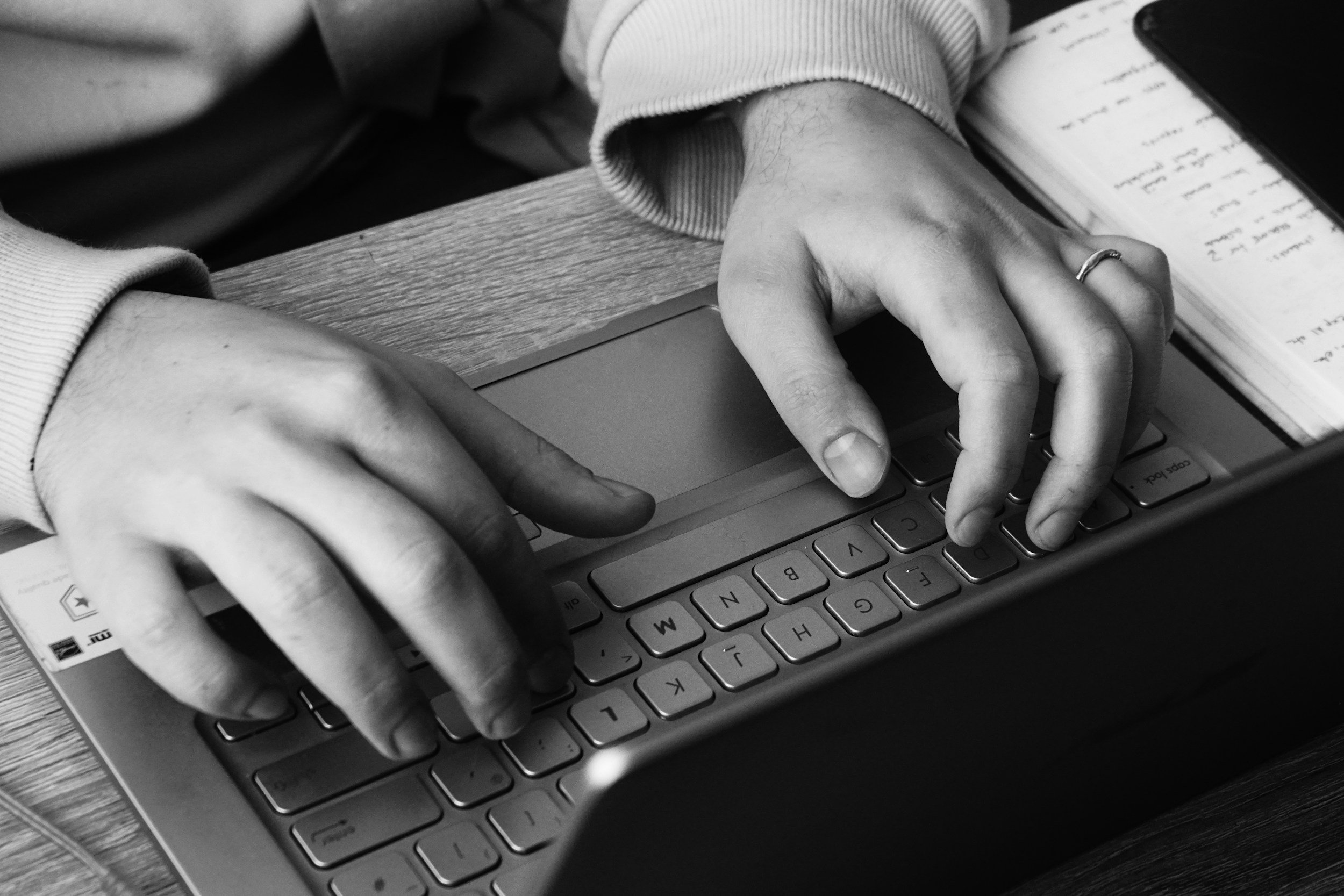 Black and white photo of a person's hands typing on a laptop keyboard with a notebook and phone nearby.