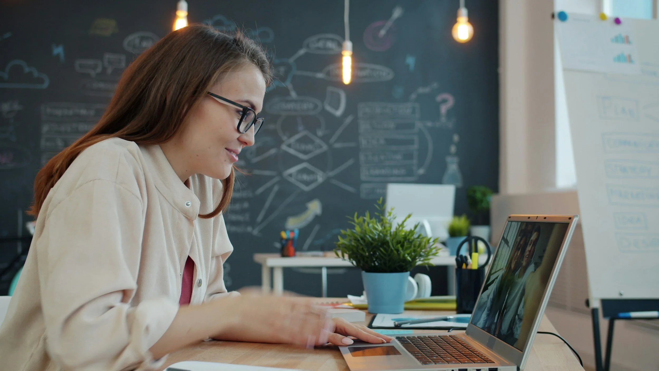 A woman sitting in front of a blackboard covered in unreadable text. She is sitting at a computer doing a call. She is wearing a casual blouse with black glasses.