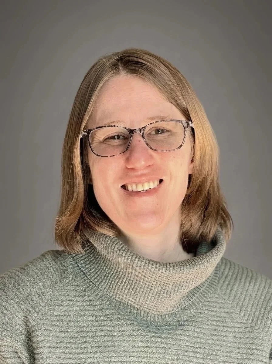 A woman with shoulder-length light brown hair, wearing glasses with a tortoiseshell frame and a gray turtleneck sweater, smiling against a gray background.
