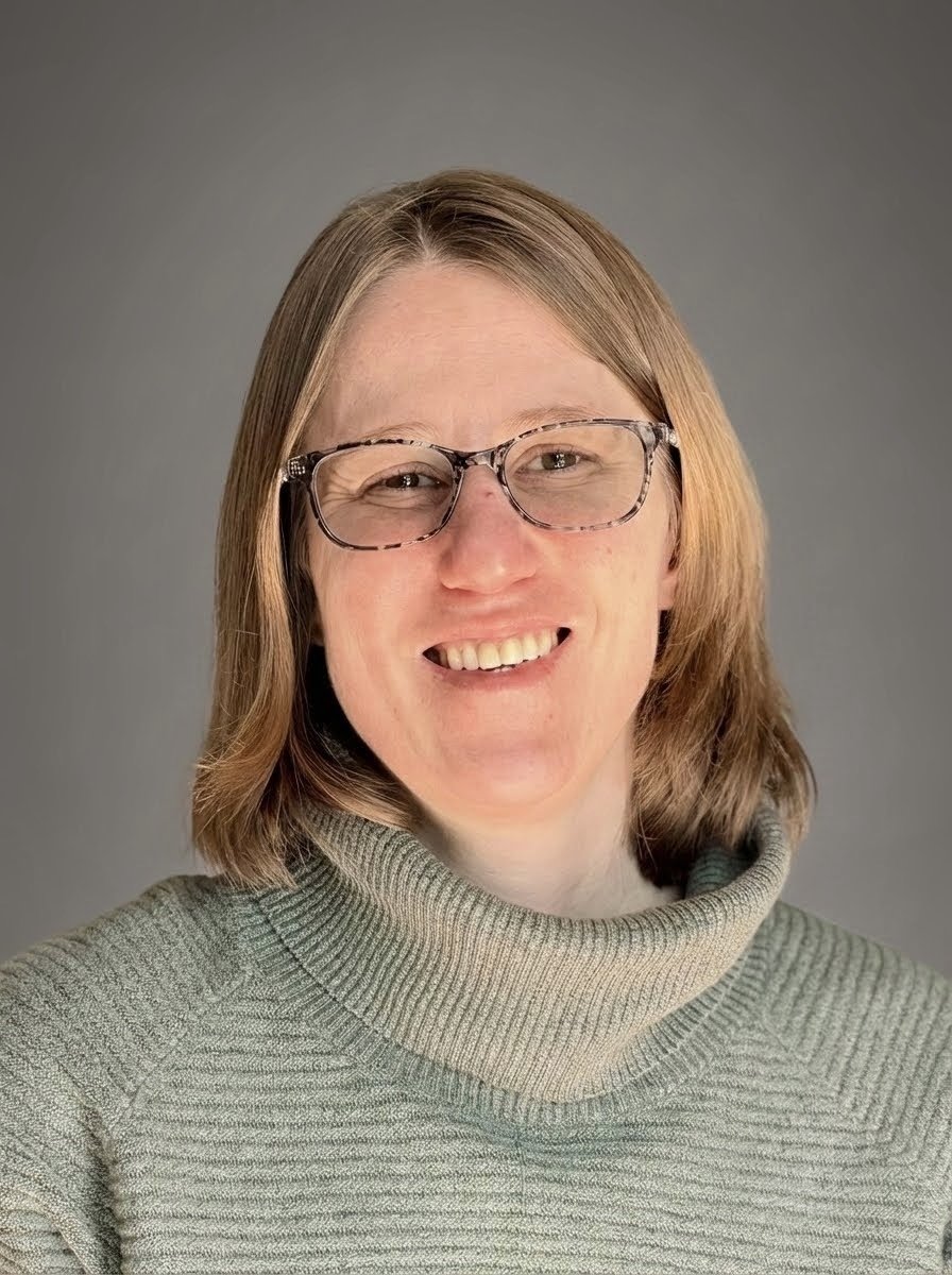 A woman with shoulder-length light brown hair, glasses, and a green turtleneck sweater, smiling at the camera against a gray background.