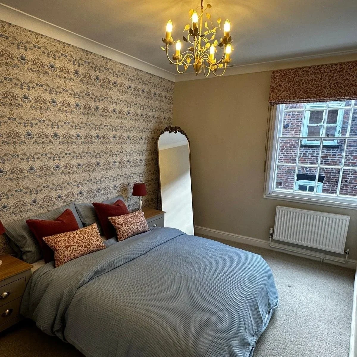 A cozy bedroom with a bed covered in grey bedding and decorated with red and patterned pillows. There is a wooden nightstand with a red lamp, a tall ornate mirror, a window showing a brick building outside, and a chandelier hanging from the ceiling. The wall behind the bed has patterned wallpaper, and the room has a carpeted floor.