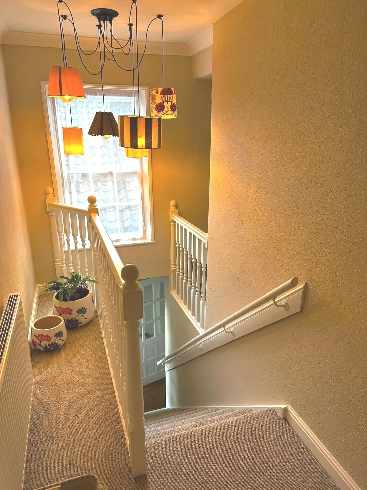 View of a staircase landing in a house with beige walls, a window, and a chandelier with multiple hanging lampshades of different colors and patterns. There are two decorative pots with plants near the bottom left corner.