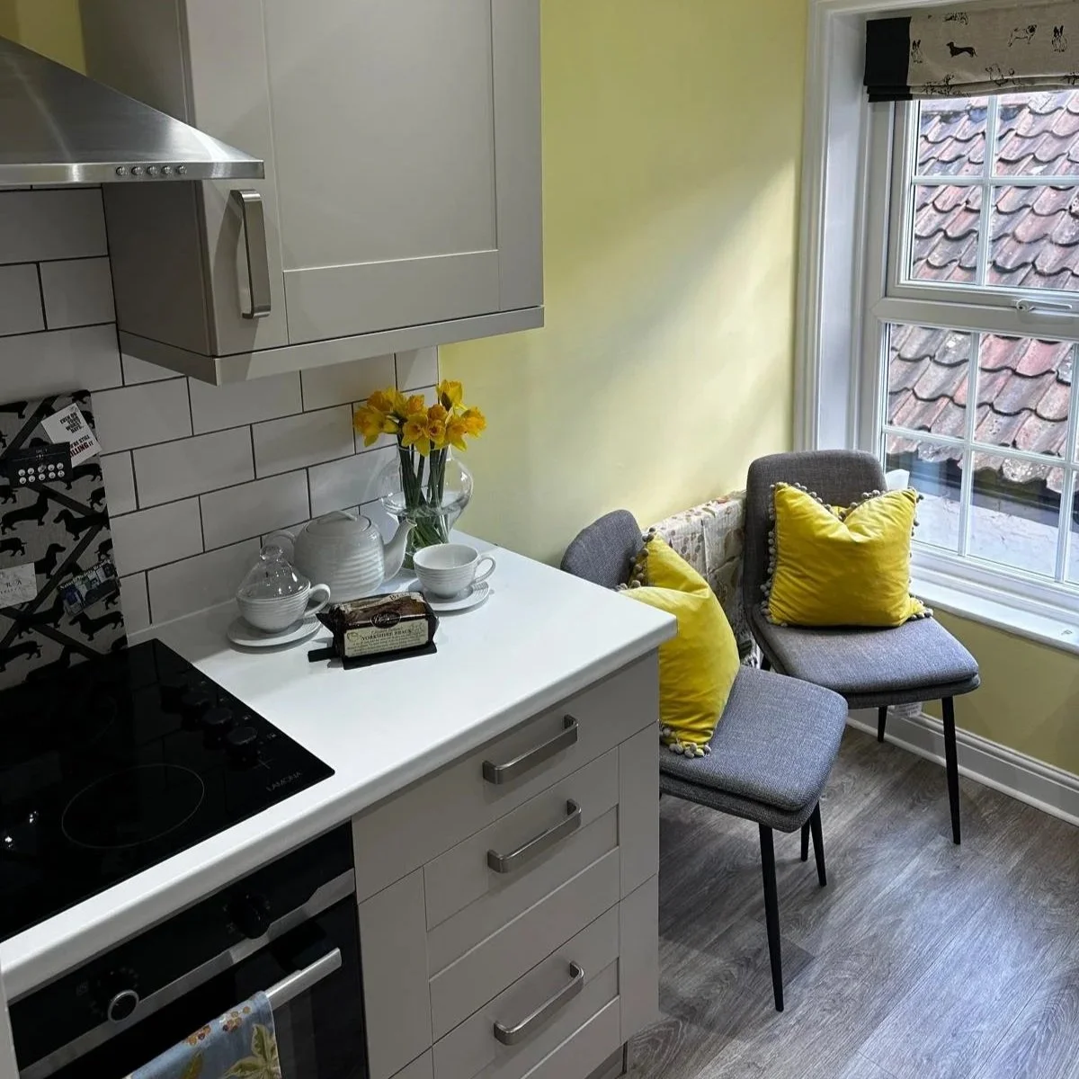 A kitchen with white cabinets, a black electric stove, a stainless steel range hood, white subway tile backsplash, and a white countertop. On the counter are a vase of yellow daffodils, a teapot, a small bowl, and some packaged snacks. There are two gray chairs with yellow pillows near a large window with a black and white dog patterned Roman shade, and a yellow wall in the background.