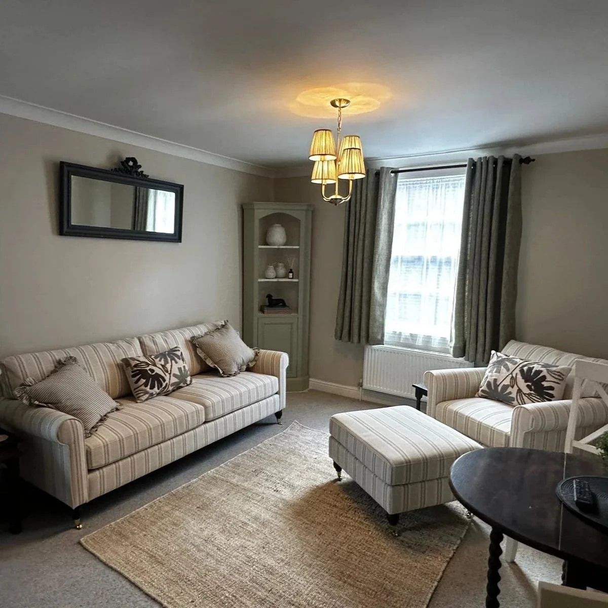 Living room with striped beige sofa, armchair with matching ottoman, beige area rug, light gray walls, window with patterned curtains, green corner cabinet, rectangular mirror, yellow chandelier, and decorative pillows.