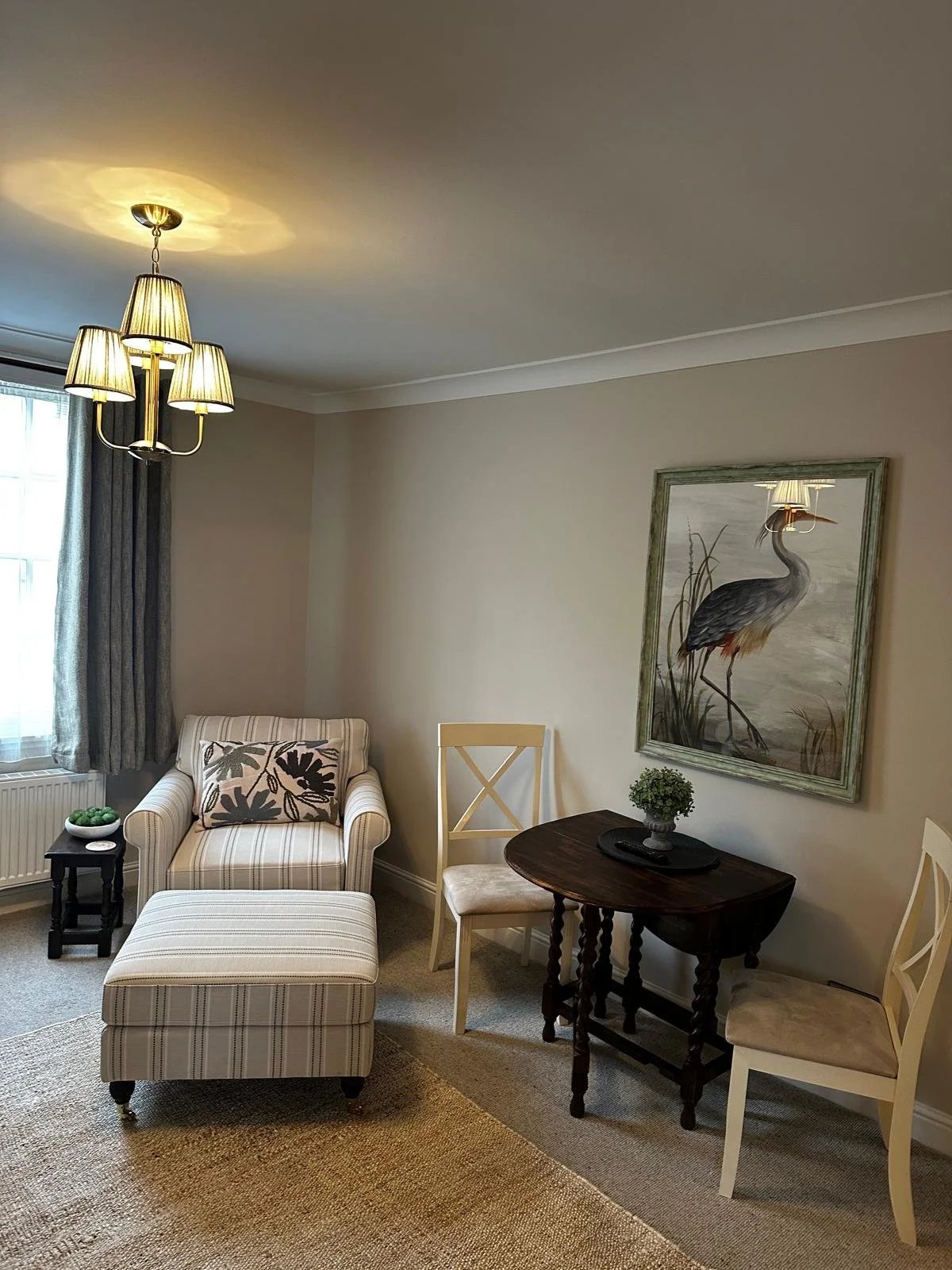 Living room with an armchair, ottoman, side table, and wall art of a heron with a lampshade reflection.