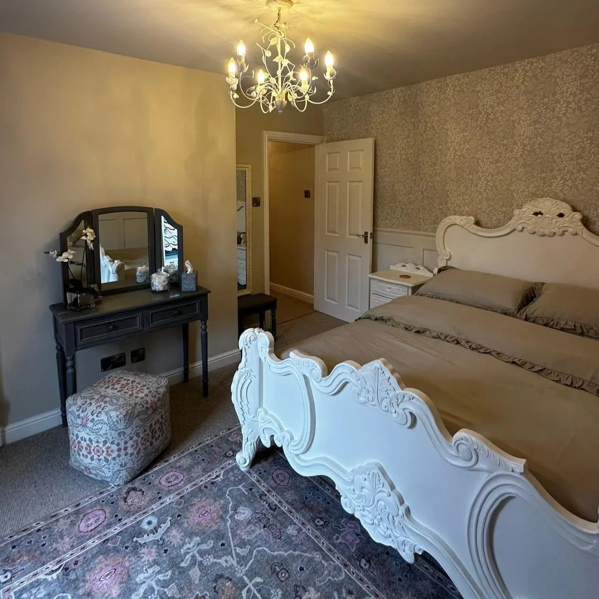 A bedroom with white ornate bed frame, beige bedding, vintage-style white nightstand, black vanity with mirror, fabric-covered stool, floral arrangement, dresser, and chandelier lighting, with floral and patterned wallpaper and a rug on the floor.