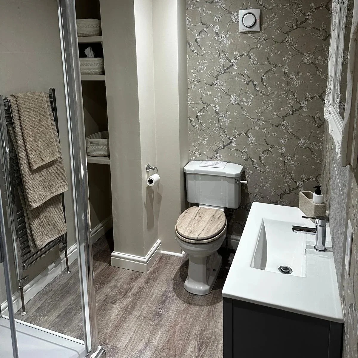 A small bathroom with wood-look flooring, a white toilet with a wooden seat, a white sink with a modern faucet, a soap dispenser, a mirror, a floral wallpapered accent wall, a towel rack with two beige towels, and a narrow open closet with baskets.