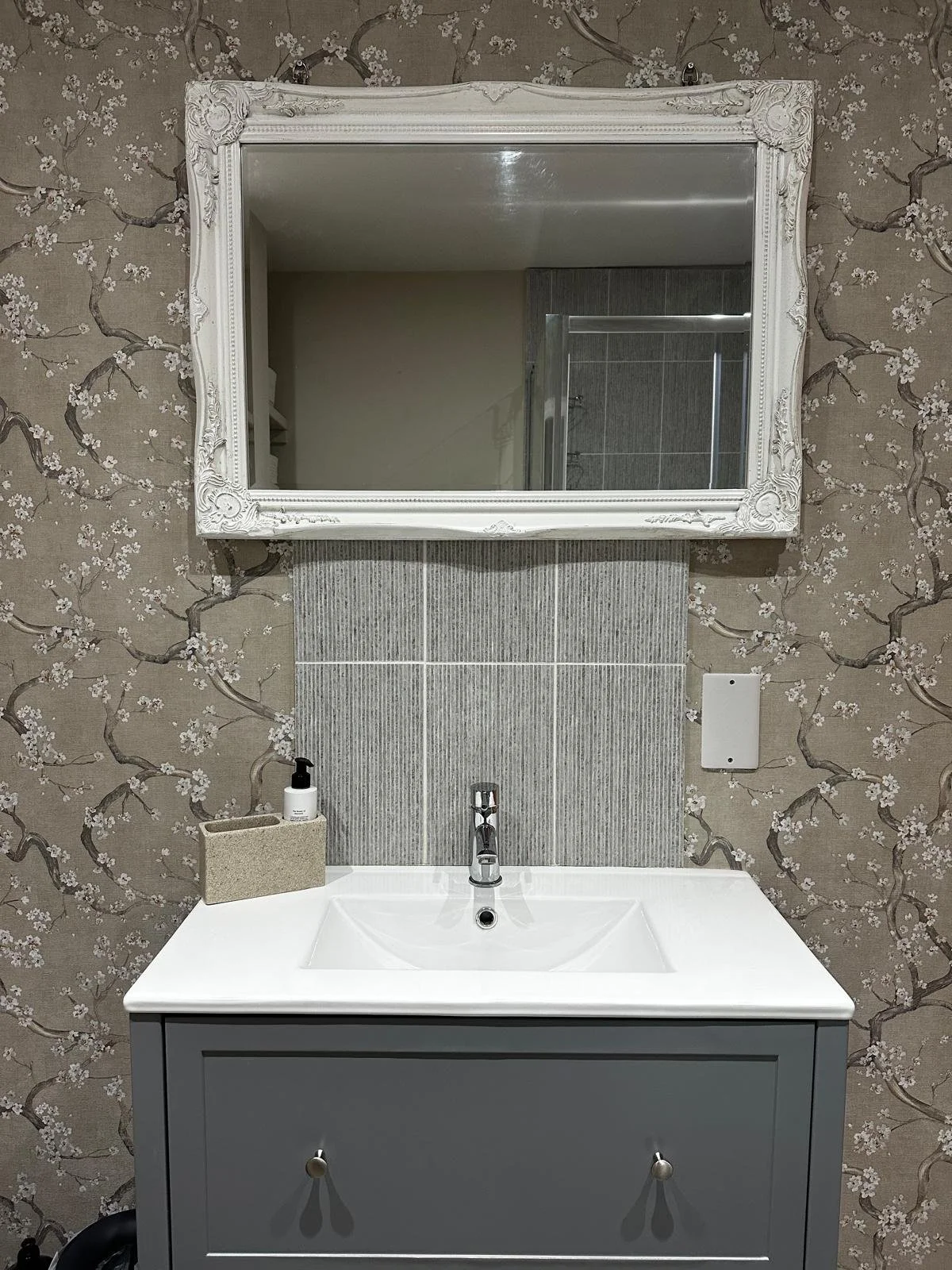 Bathroom sink with a gray cabinet, white countertop, and a rectangular mirror with ornate white frame mounted on floral wallpapered wall. Items on the counter include a soap dispenser and a toothbrush holder. The reflection in the mirror shows part of the shower area with a tiled wall.