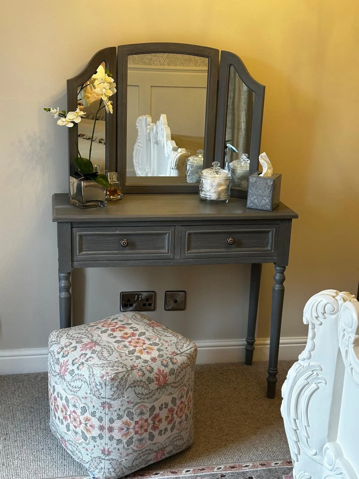 A gray wooden vanity with a tri-fold mirror, decorated with a potted orchid, glass jars, a tissue box, and perfume bottles, accompanied by a floral upholstered ottoman.