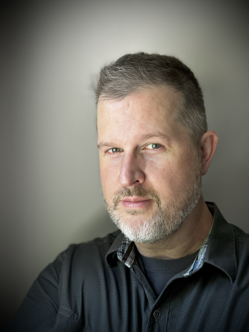 A middle-aged man with short gray hair and a beard, looking at the camera with a serious expression, wearing a black jacket over a dark shirt, against a blurred gray background.
