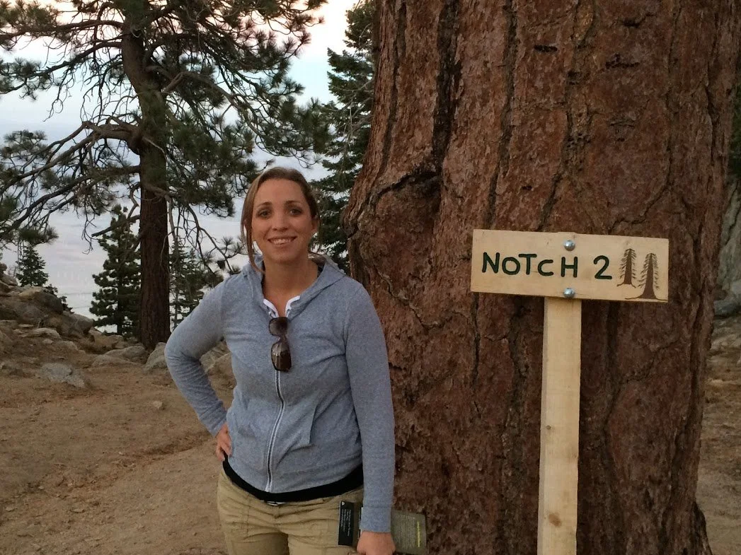 A woman standing next to a large pine tree with a sign that reads "NOTCH 2" with drawings of trees. The woman is wearing a gray zip-up jacket, khaki pants, and sunglasses hanging from her jacket. The background features additional pine trees and a cloudy sky.