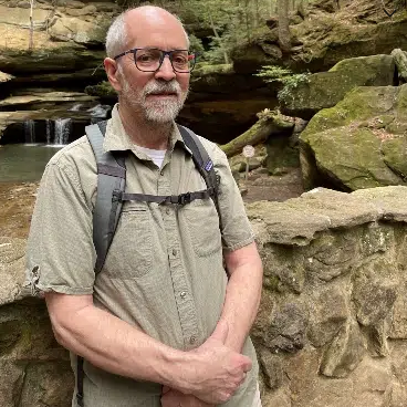 An older man with glasses, a beard, and short gray hair, wearing a light khaki short-sleeve shirt and a backpack, standing outdoors near a rocky stream with moss-covered rocks and a small waterfall.