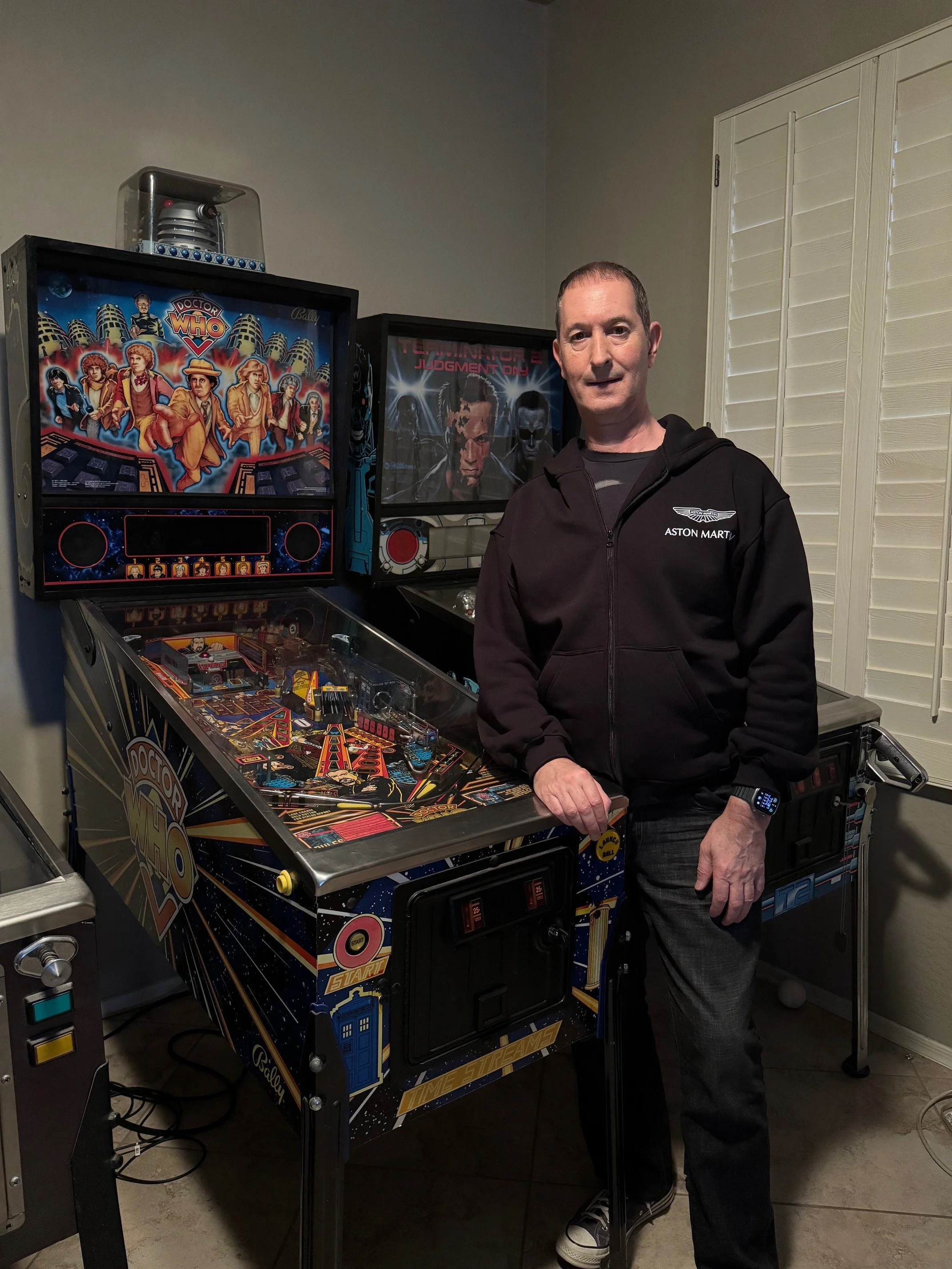 A man standing next to a vintage Doctor Who-themed pinball machine in a room with beige tiled flooring and cream-colored wall. The man is wearing a black Aston Martin hoodie and jeans.