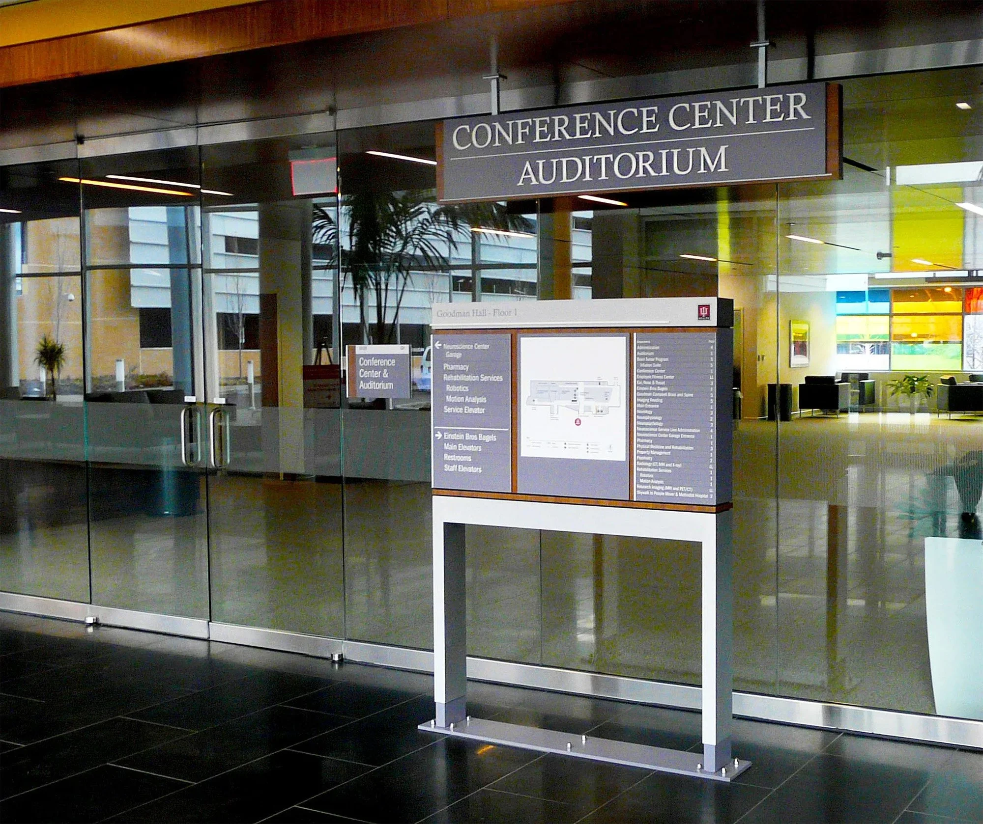 Inside view of a conference center lobby with a directory sign and glass walls. The sign reads "Conference Center Auditorium" and lists various facilities and directions nearby.