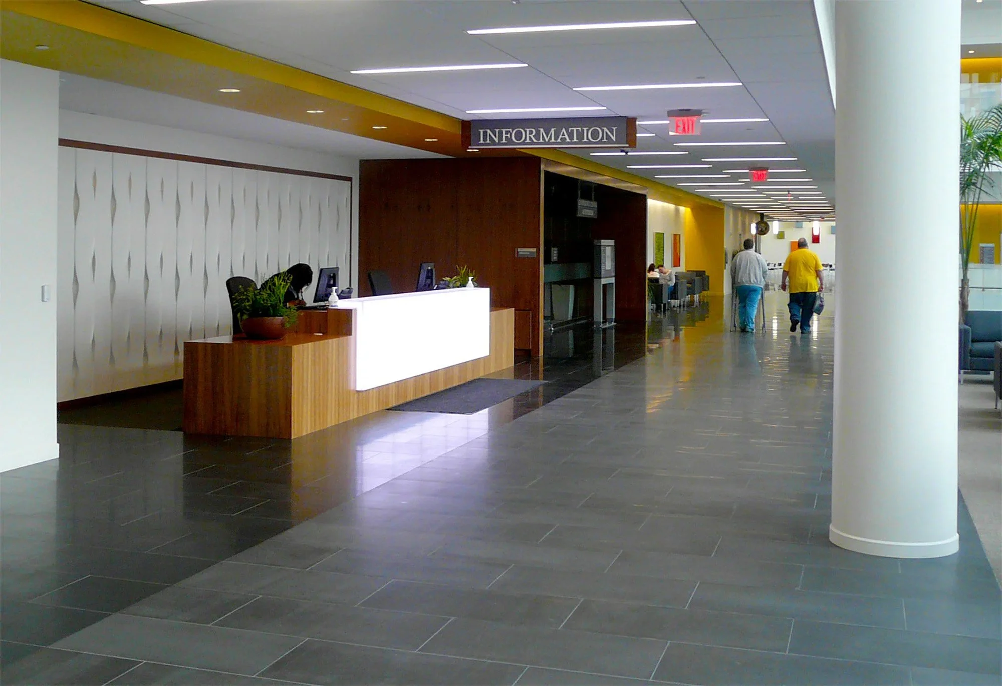 A modern hospital or office building interior with a reception desk labeled "Information," a large white column, a yellow accent wall, and a hallway with people walking.