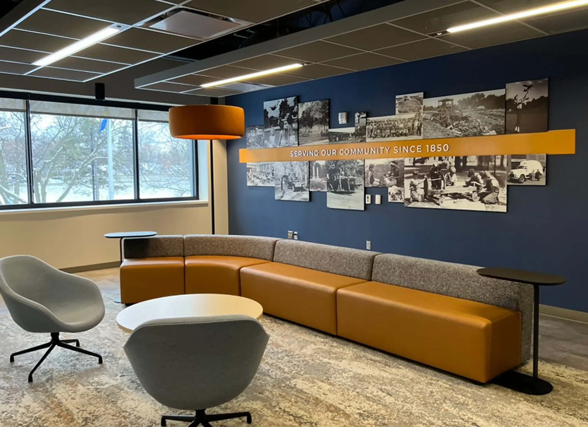 Modern community waiting area with curved orange and gray sofa, two gray chairs, a white oval coffee table, a large window showing snow-covered trees outside, and a navy blue wall with historical black-and-white photographs and a yellow banner that reads 'Serving Our Community Since 1850.'