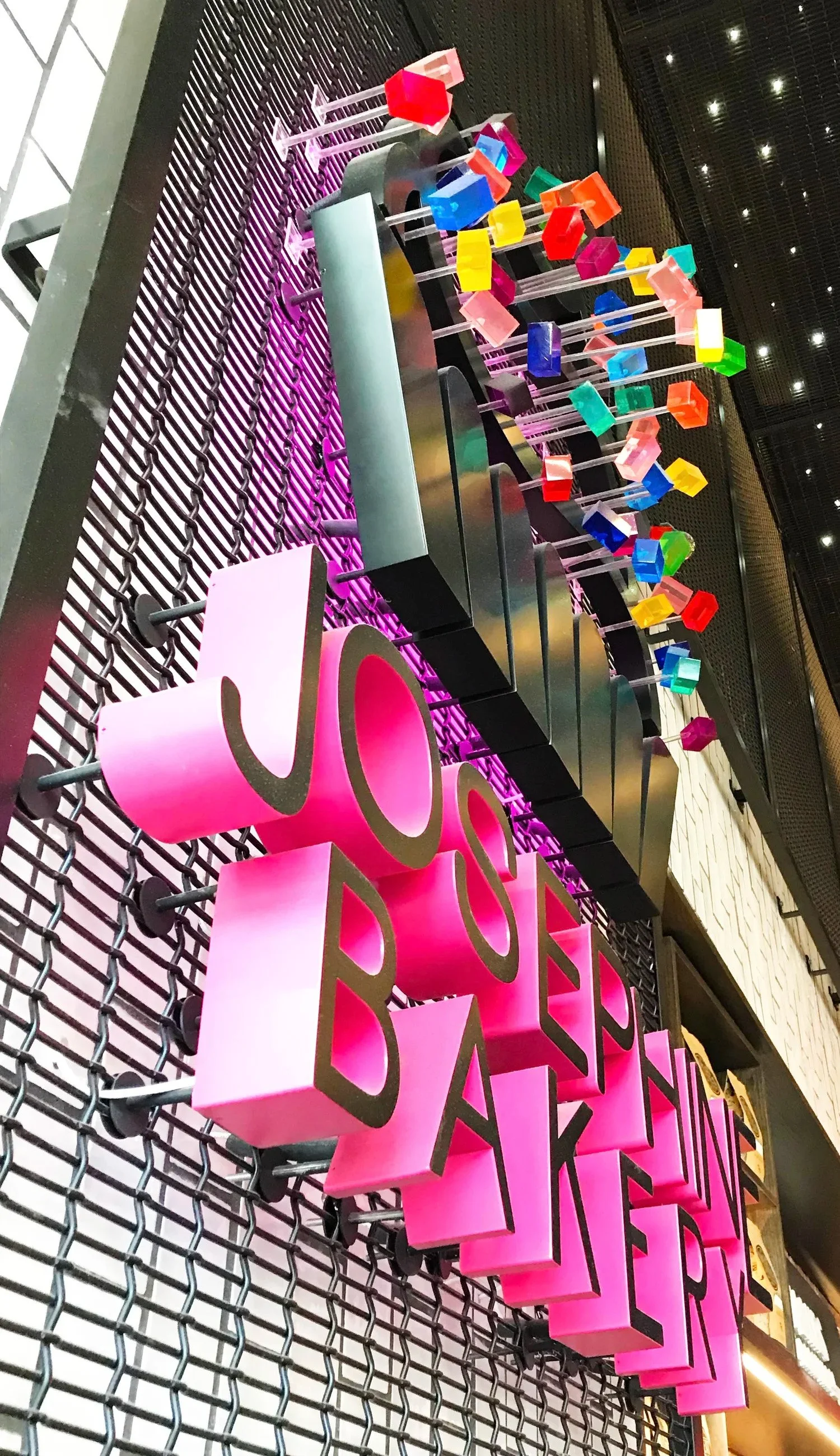 Colorful illuminated sign with the words 'JOSEPH BAKERY' and decorative colorful cubes on rods, mounted on a black metal grid.